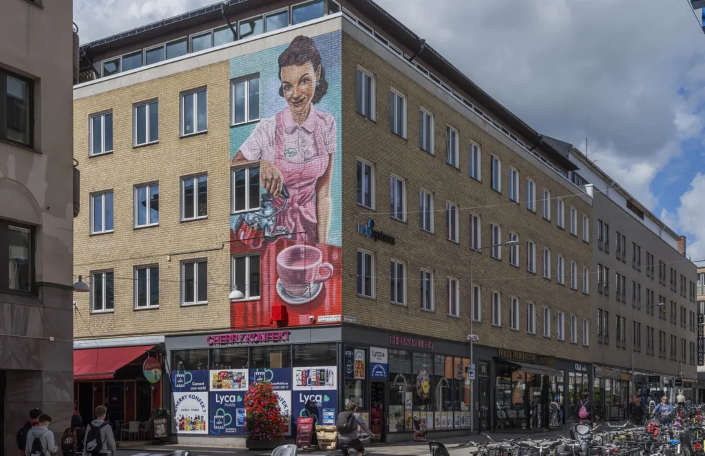 En gul byggnad med en stor väggmålning av en kvinna som håller en kaffekanna, samt butiker på bottenvåningen.