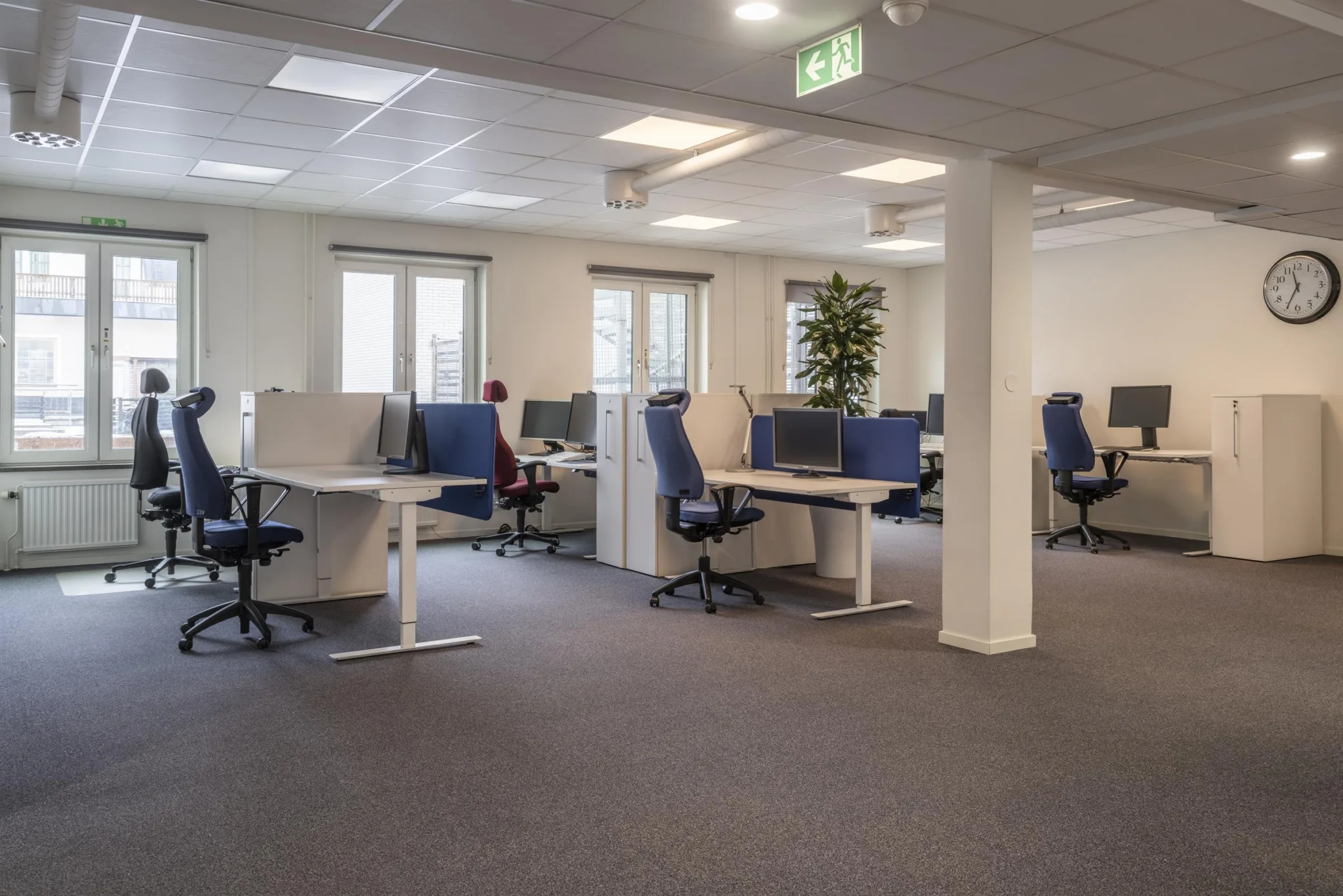 En öppen kontorsmiljö med flera arbetsstationer, stolar och skrivbord, ljusa väggar och stora fönster som släpper in naturligt ljus.
