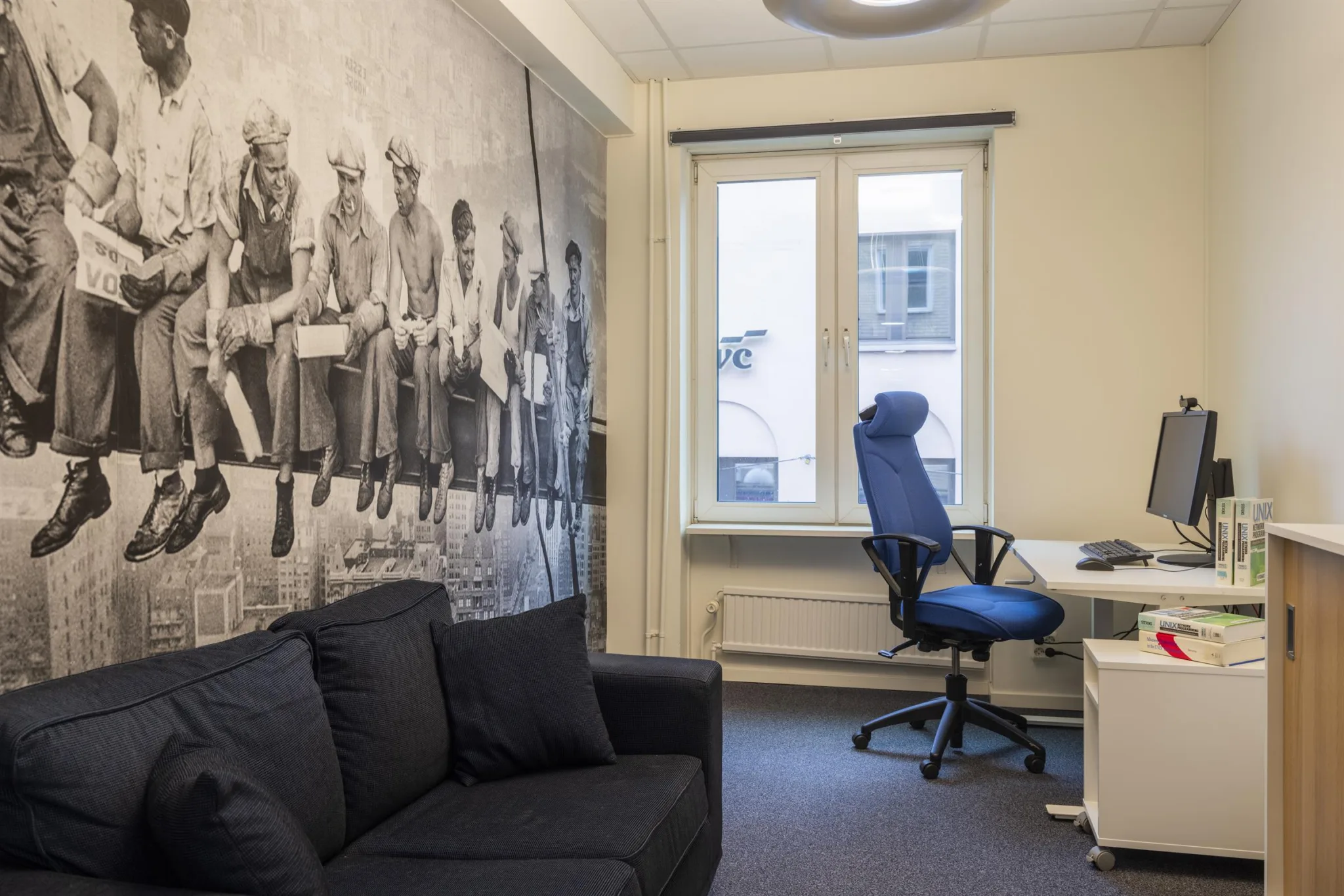 Ett kontorsrum med en svart soffa, en skrivbordsstol vid ett skrivbord och en vägg med en stor svartvit väggmålning av byggnadsarbetare som sitter på en balk.