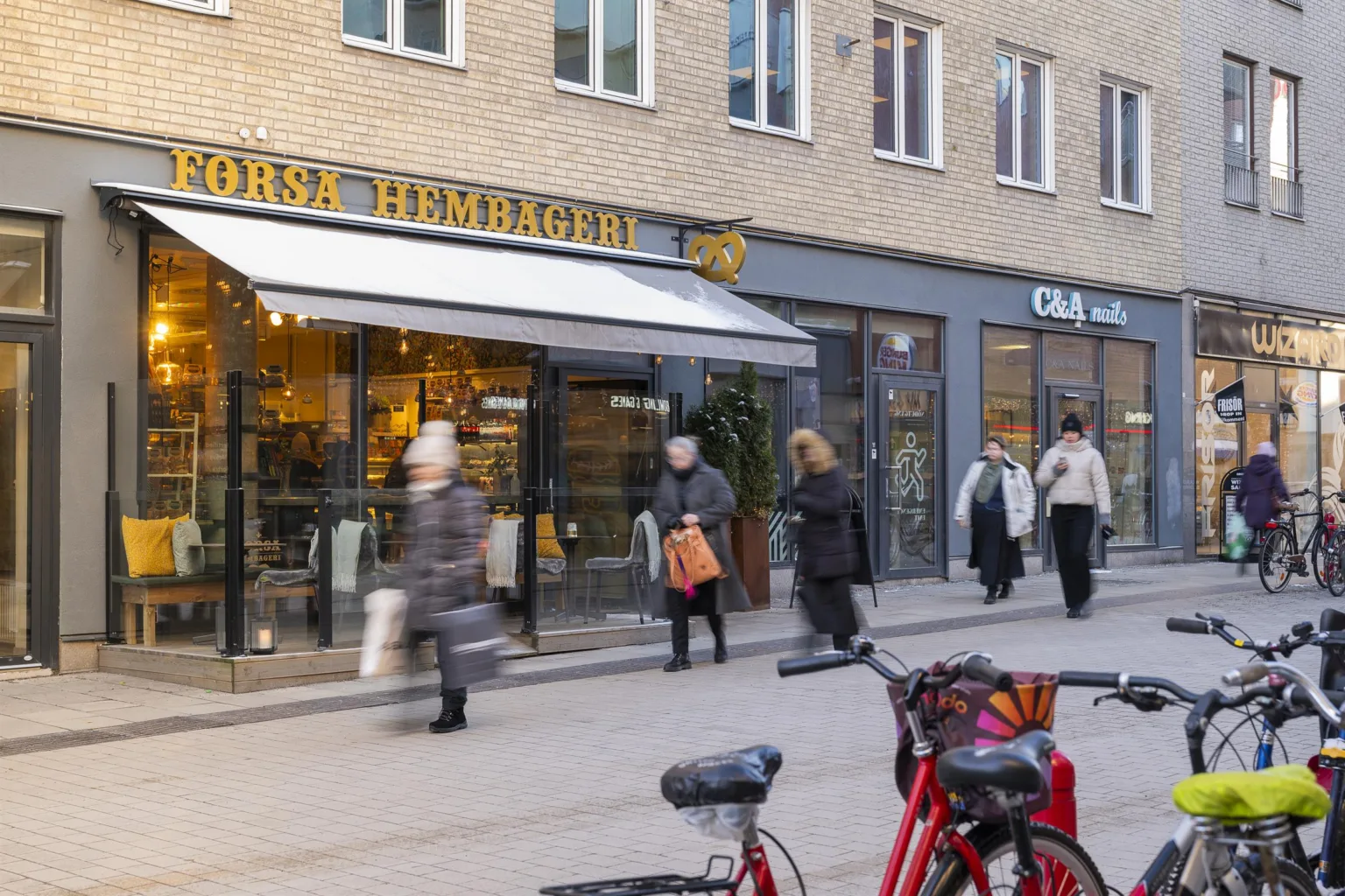 En gatuvy med människor som passerar framför skyltfönster, inklusive en butik med skylten "Örsa Hemköp," cyklar parkerade i förgrunden.