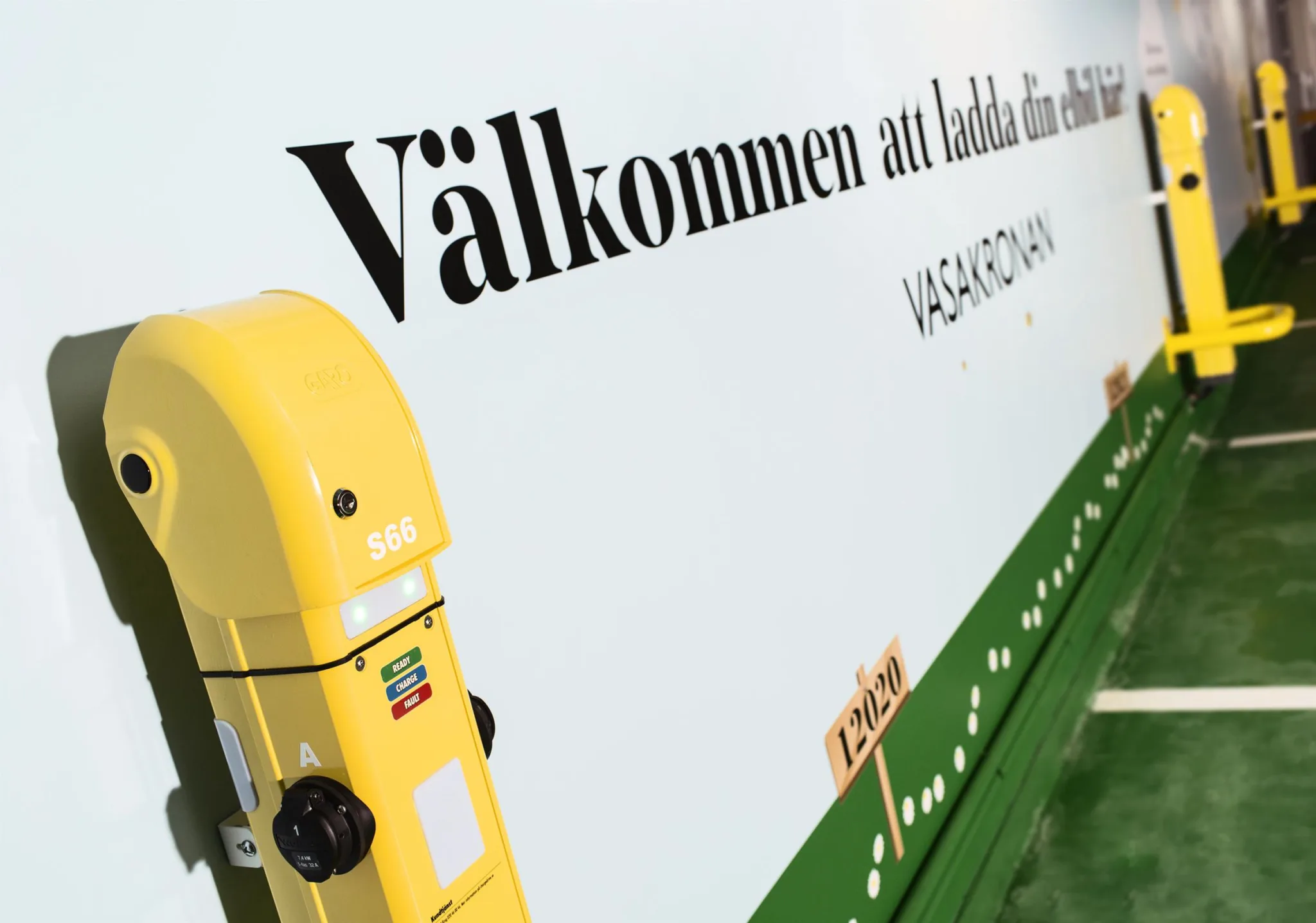 En gul elbilsladdare framför en vägg med texten "Välkommen att ladda din elbil. Vasakronan".