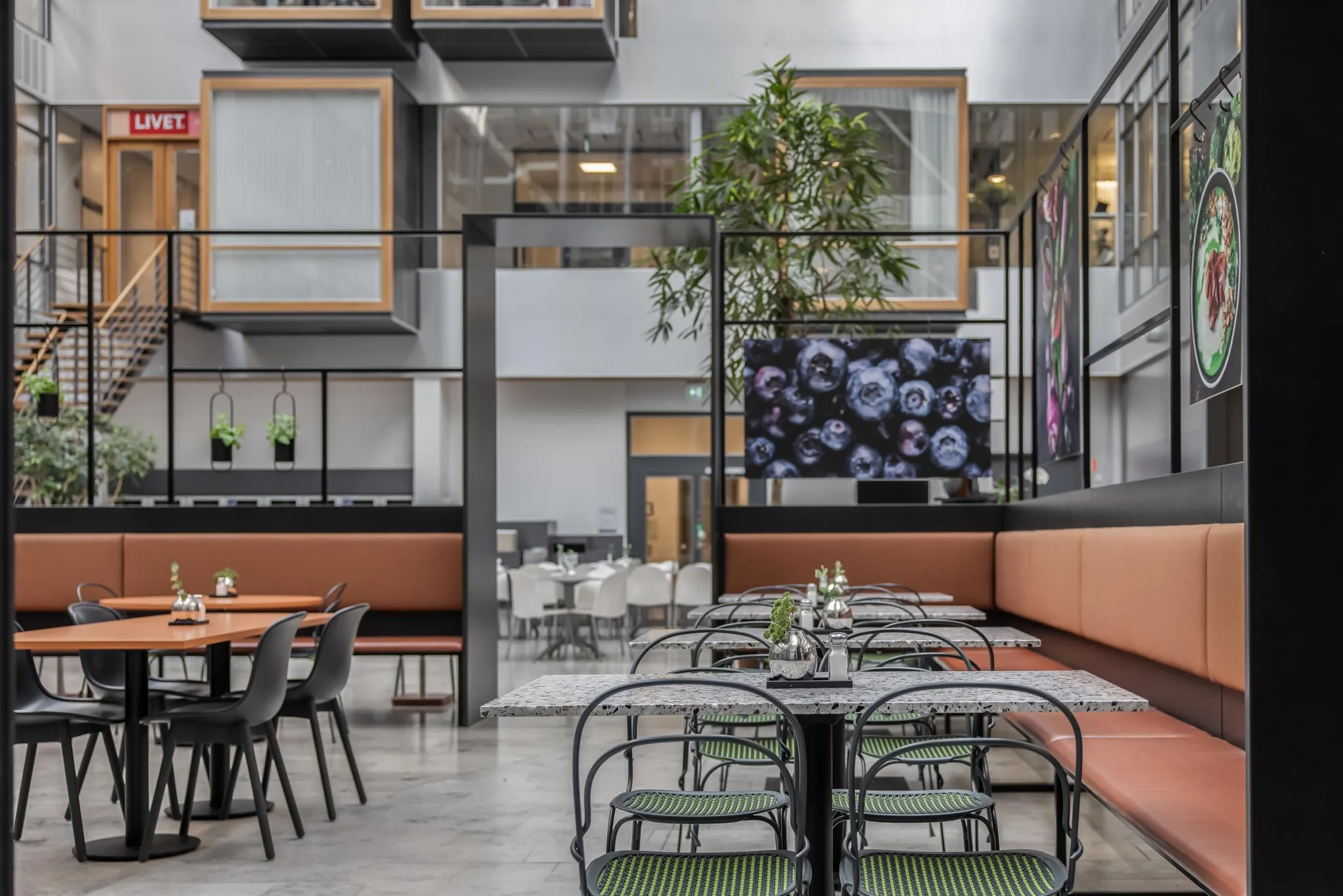 En rymlig restaurang med bord och stolar, grön inredning, växter och stora dekorativa fönster.