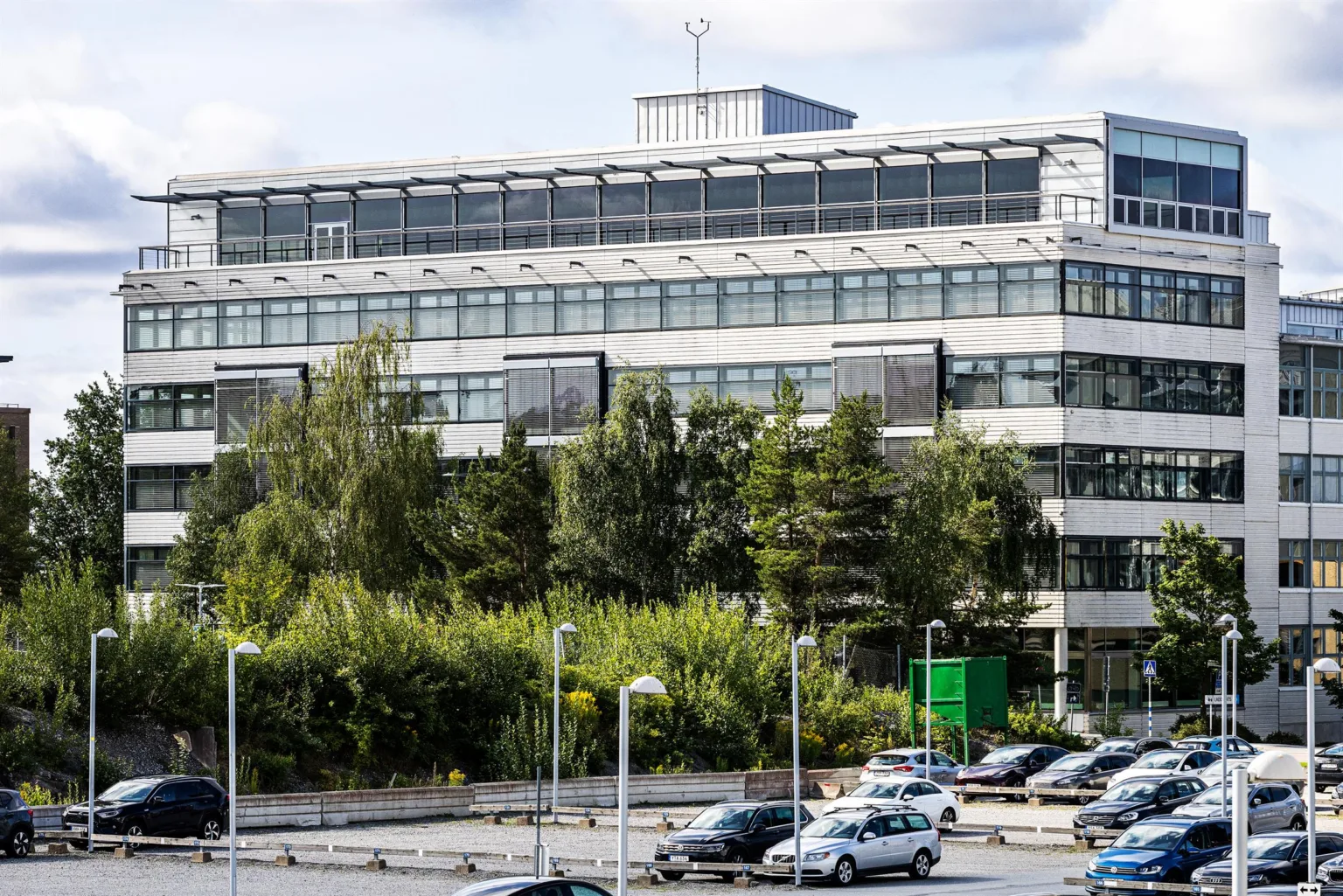 En modern byggnad med glasfasad, omgiven av träd och parkeringsplatser.