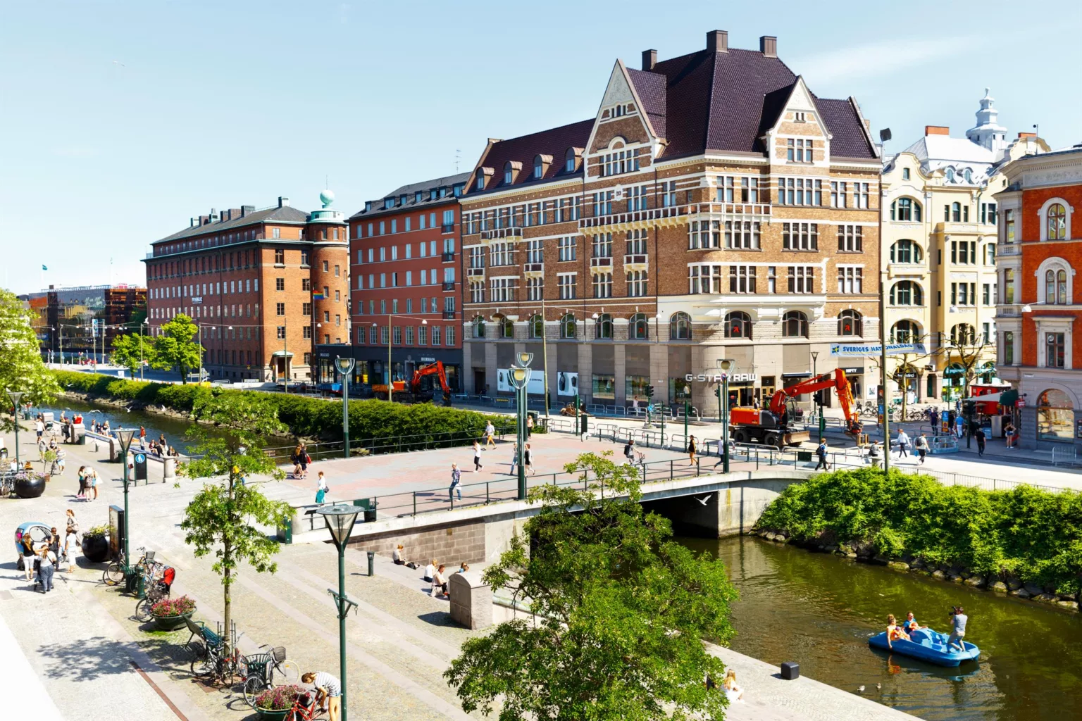 1. Flera byggnader i klassisk arkitektur vid en kanal med bro och grönska, människor promenerar på torget.