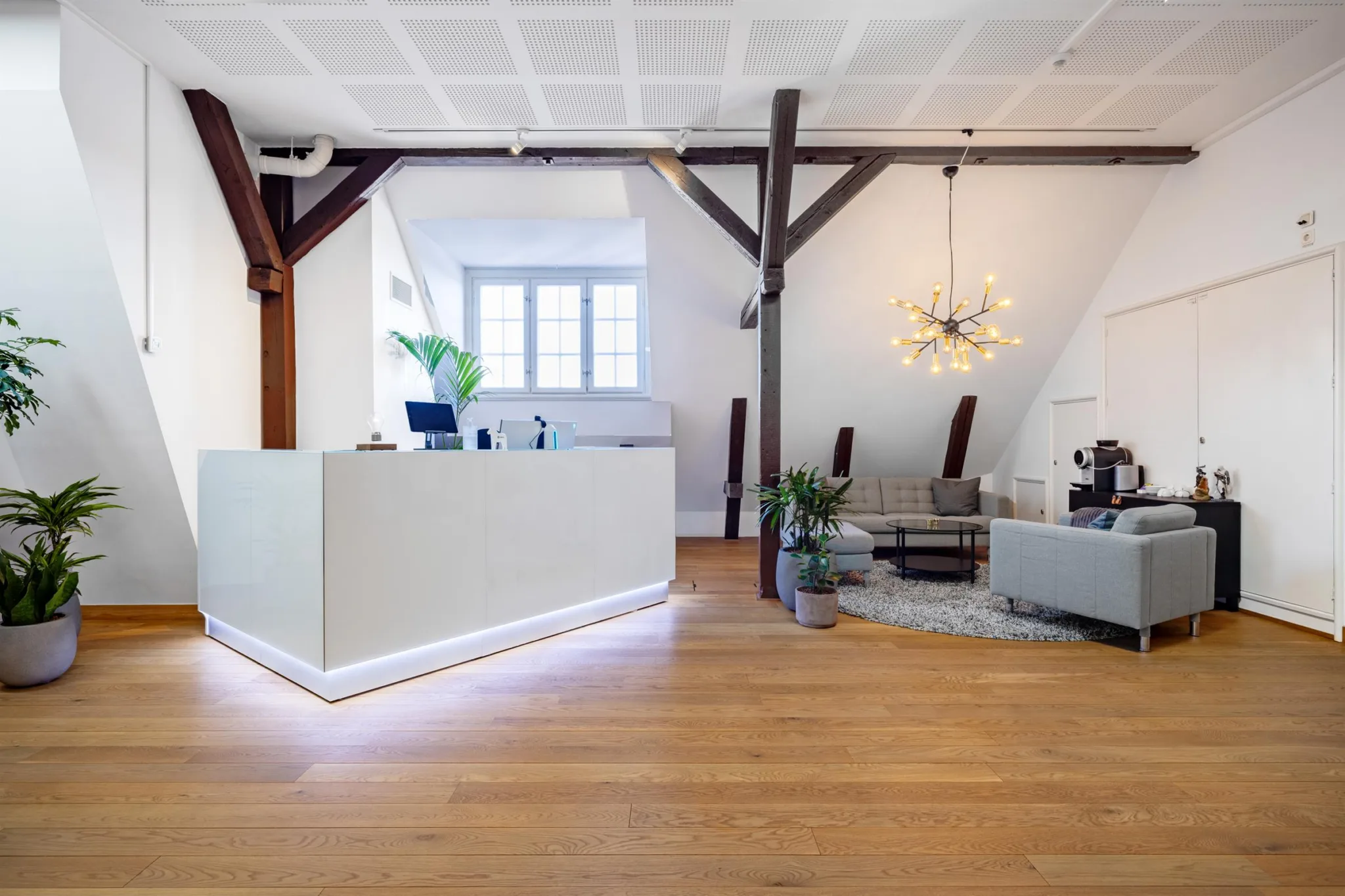 En reception med ett vitt skrivbord, en sittgrupp, en designlampa och synliga träbjälkar i taket.