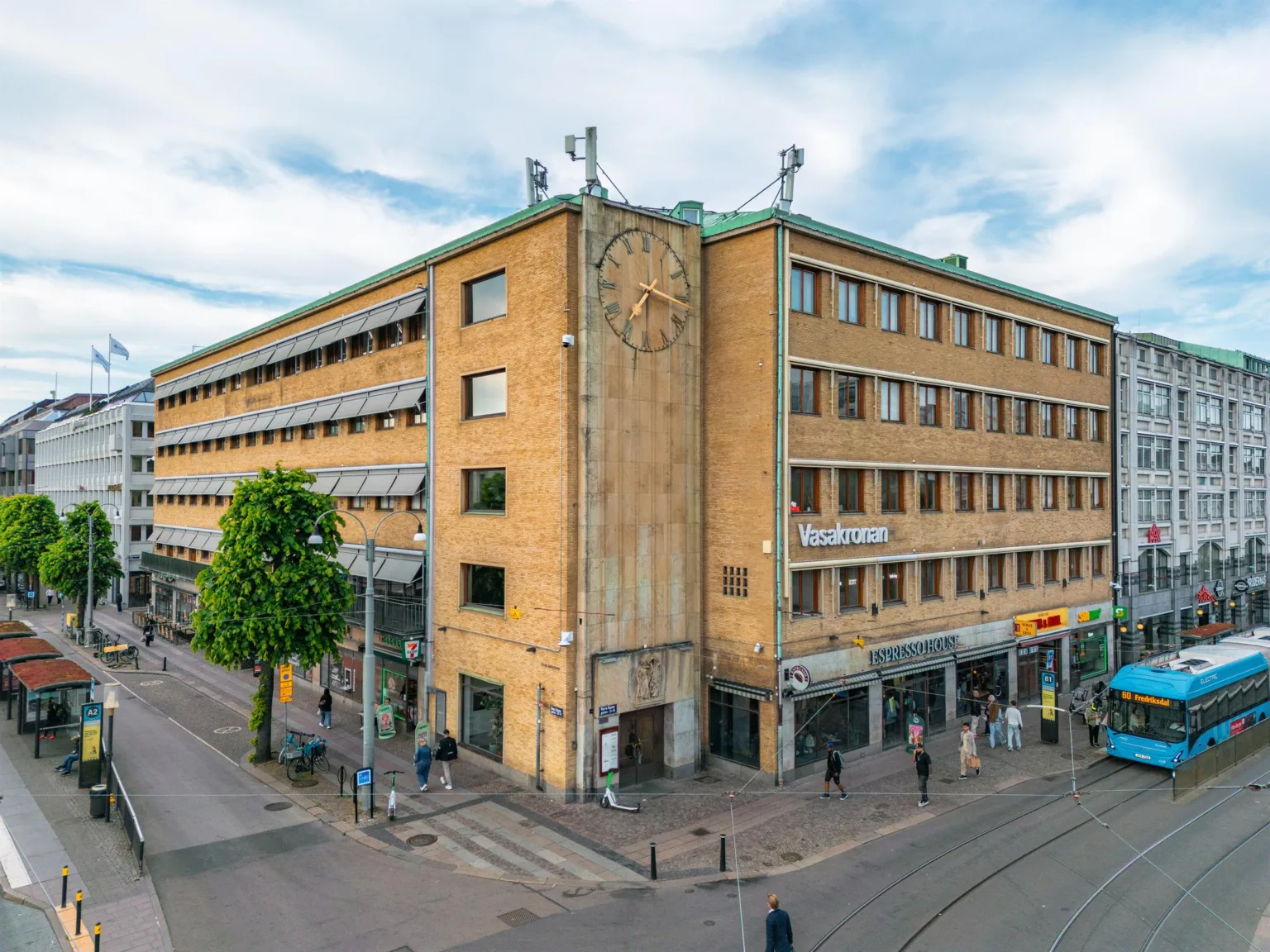 Ett hörnhus i gul tegel med en stor klocka på fasaden, omgivet av trottoarer och människor.
