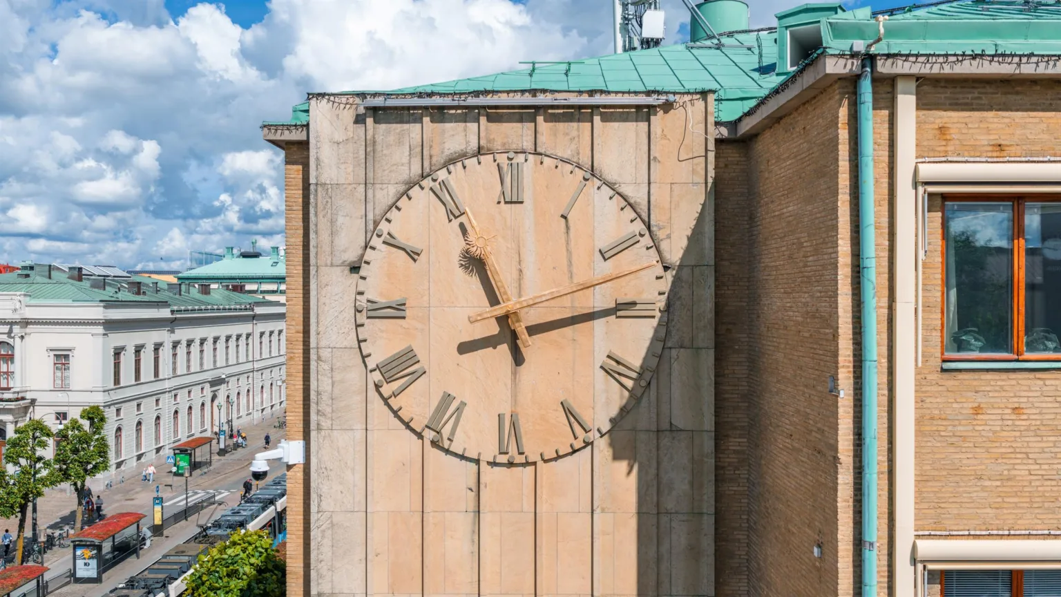 En stor klocka med romerska siffror på en beige vägg, omgiven av byggnader och en blå himmel med moln.