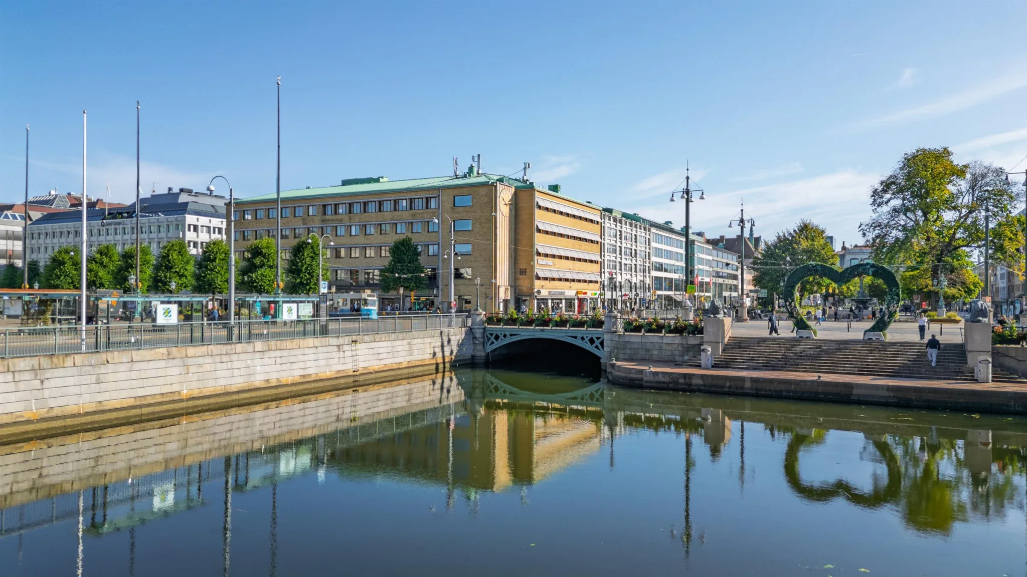 En bro över en kanal med spegelblankt vatten, omgiven av byggnader och grönska under en klarblå himmel.