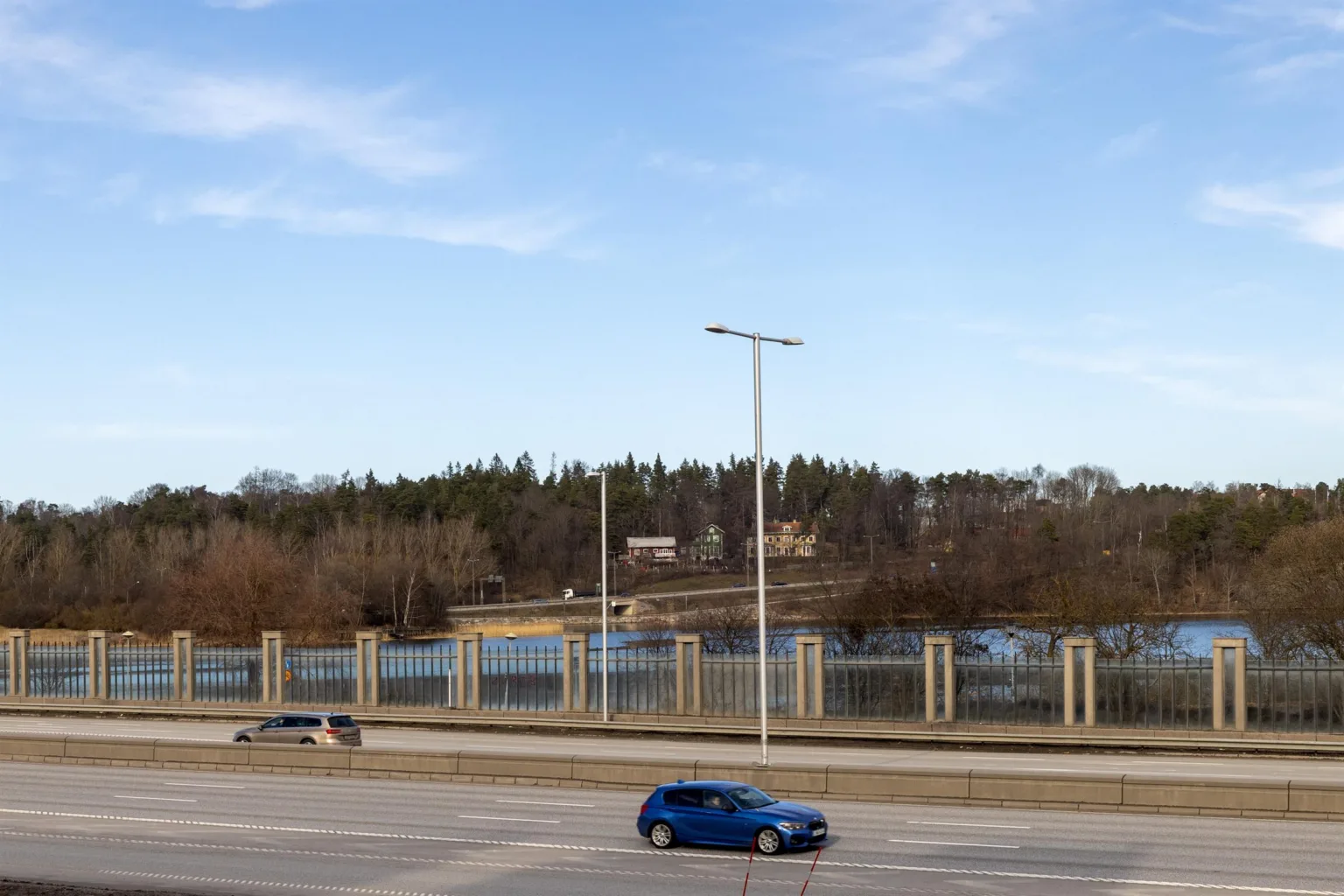 En blå bil kör på en väg med ett staket och träd i bakgrunden under en klar himmel.