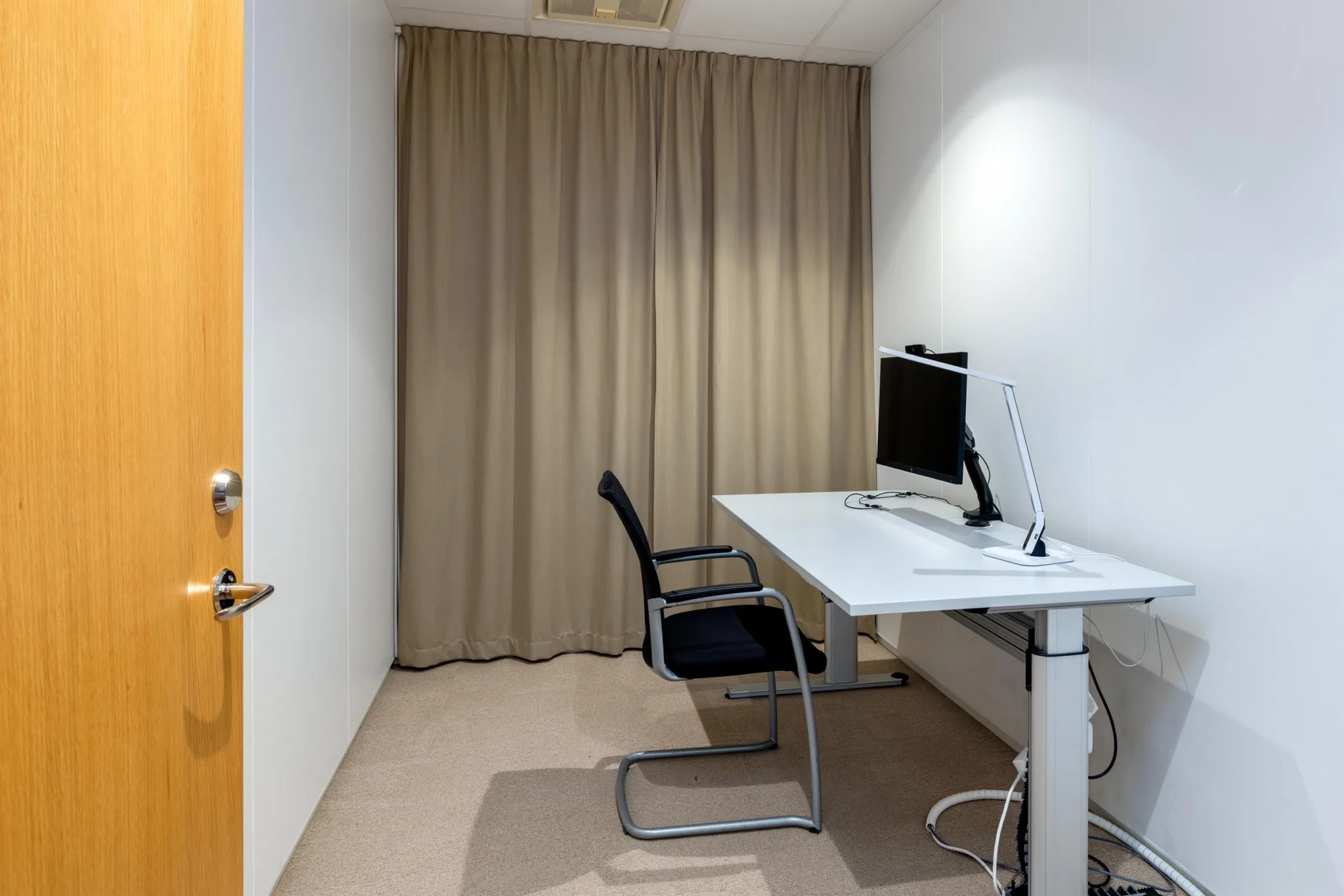Ett litet kontorsrum med ett skrivbord, en stol och en datorskärm framför beige gardiner.