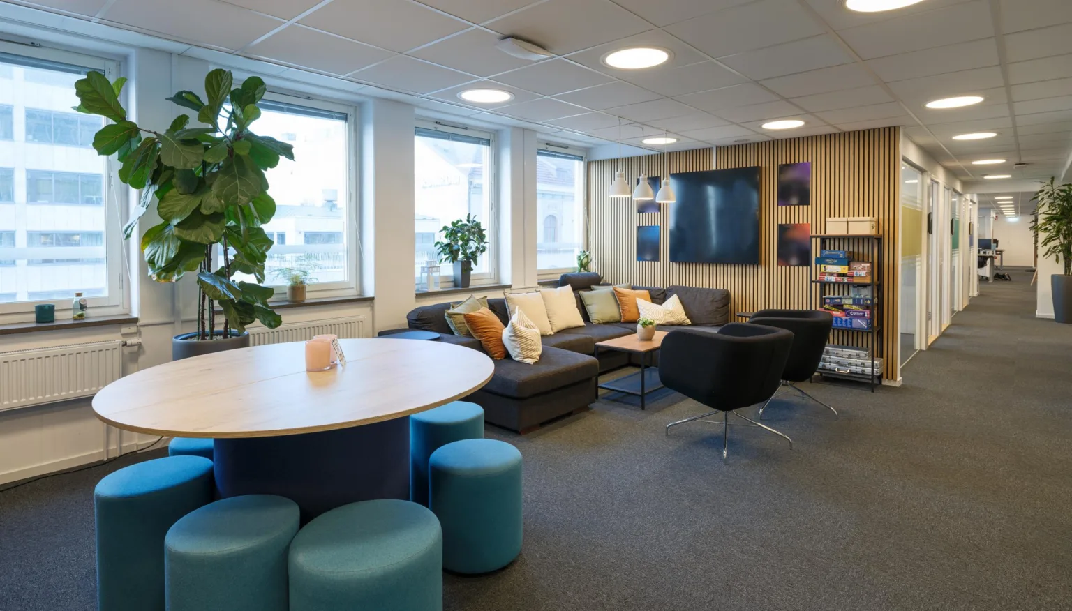 1. Ett mysigt kontorsutrymme med soffor, fåtöljer, växter och en stor skärm på väggen.