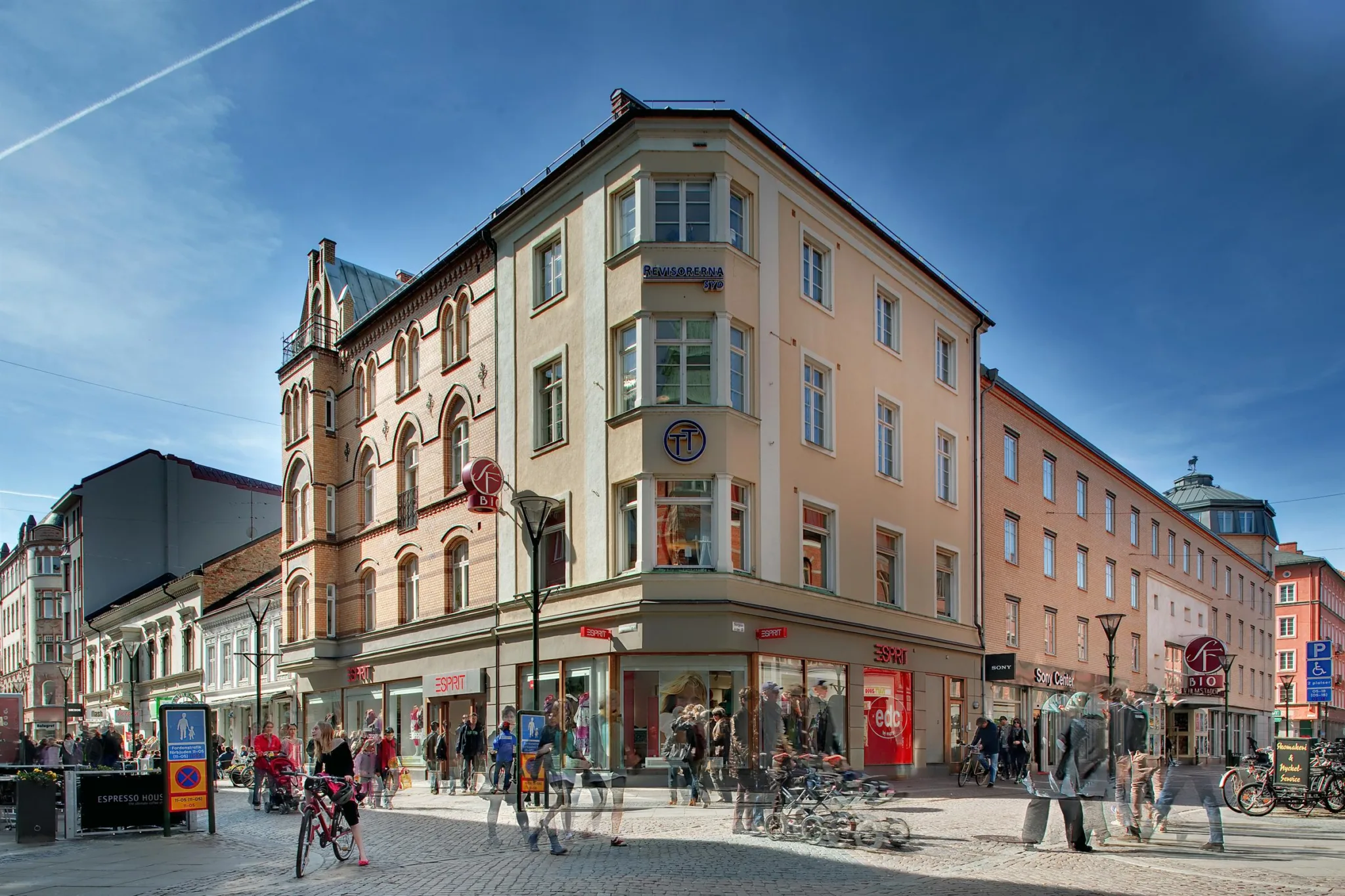 1. Ett hörnhus med flera våningar, beige fasad och butiker på bottenvåningen, omgiven av människor och cyklar på en solig dag.