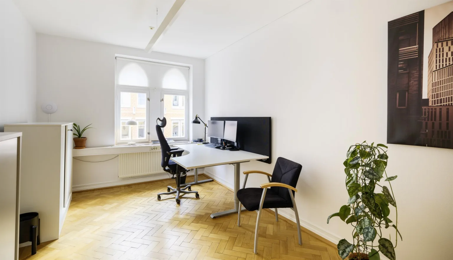 3. Ett minimalistiskt kontor med ett skrivbord, en datorskärm, en kontorsstol och en extra stol, beläget vid ett fönster med bågar.