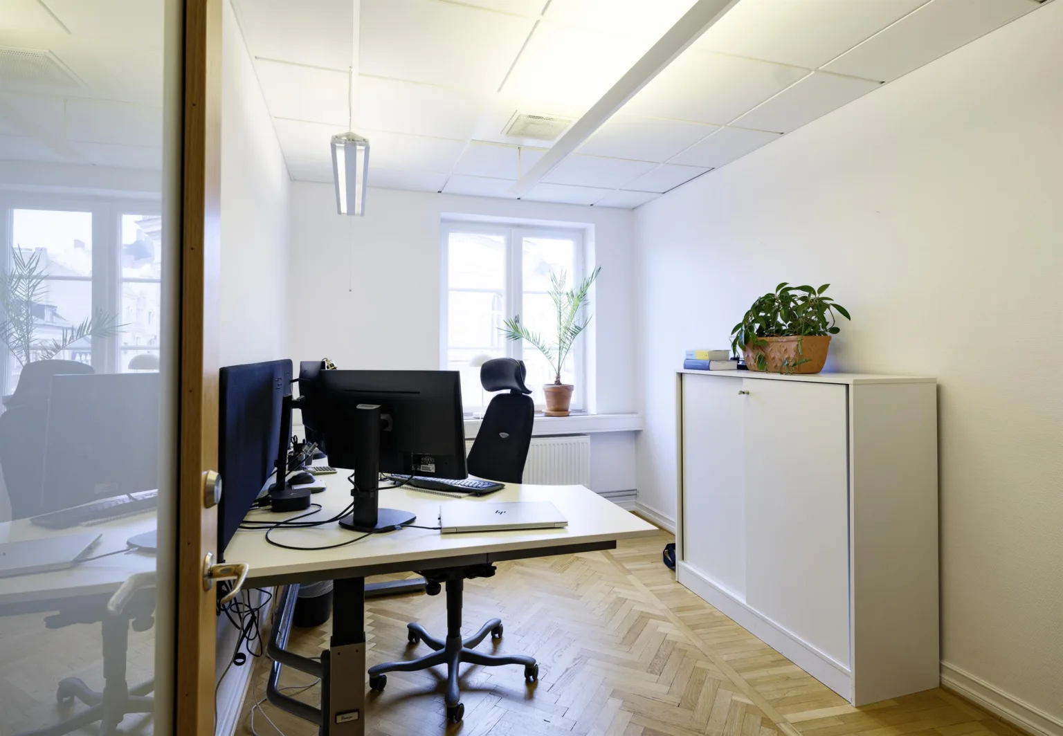2. Ett ljust kontorsrum med två skrivbord, datorskärmar, en kontorsstol och ett skåp med en grön växt.