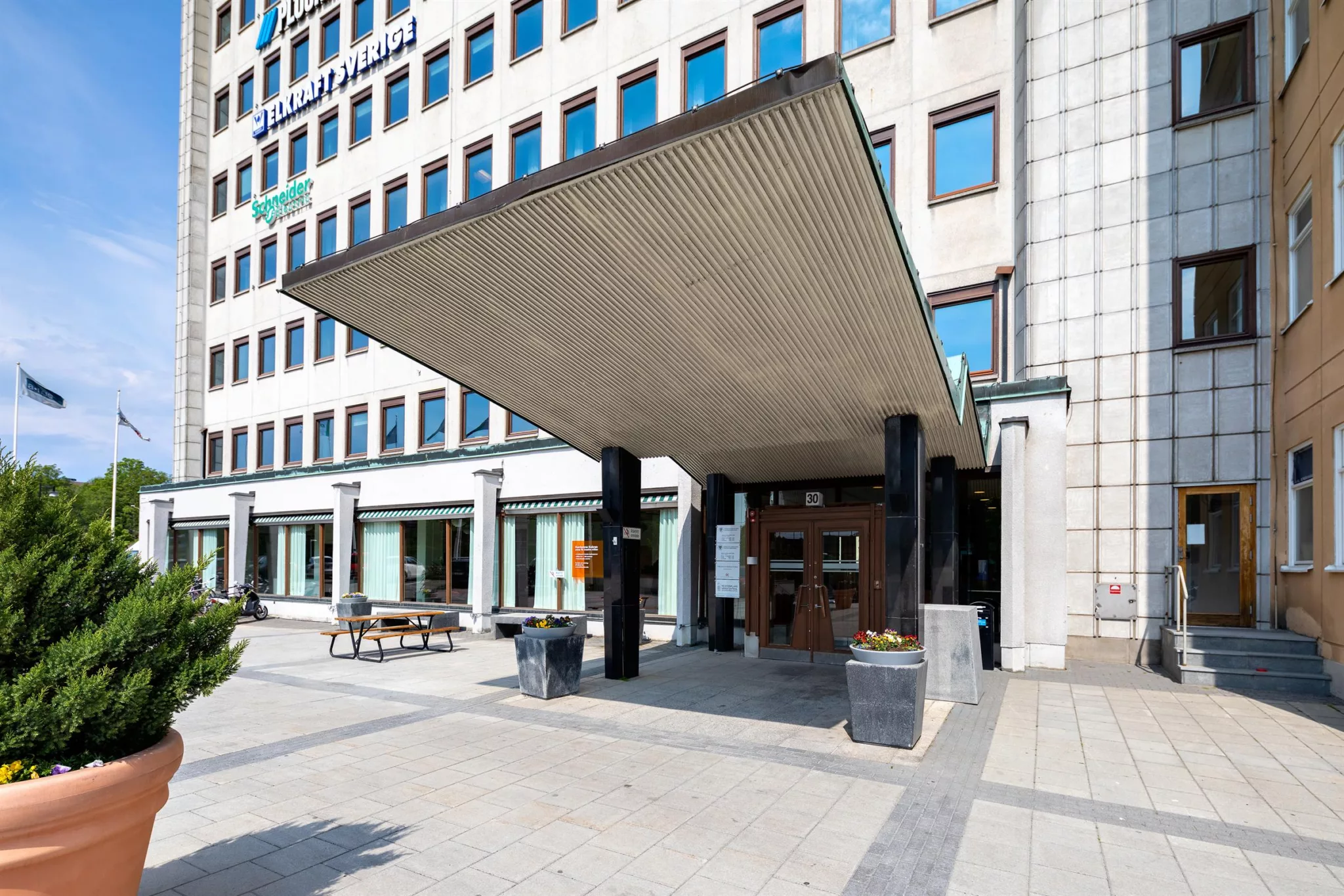 En byggnad med en stor taköverbyggnad vid entrén, omgiven av planteringskärl och bänkar.