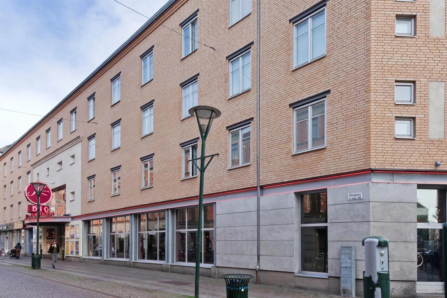 - Fyravåningshus med beige fasad och stora fönster på bottenplan, en lyktstolpe står framför byggnaden.