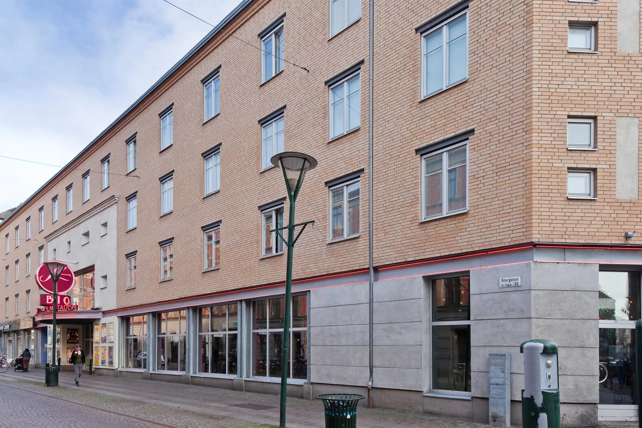 - Fyravåningshus med beige fasad och stora fönster på bottenplan, en lyktstolpe står framför byggnaden.