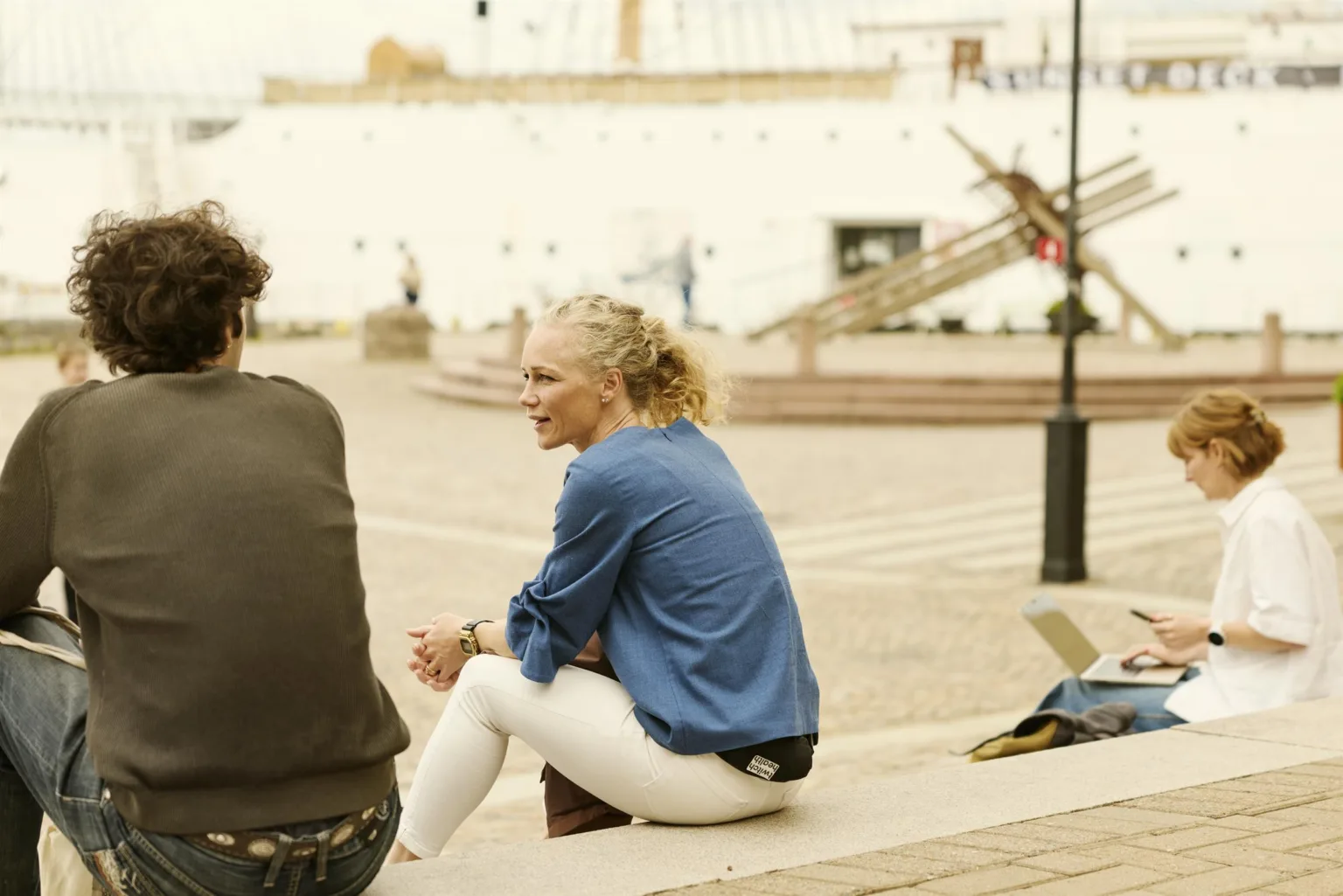 Två personer sitter utomhus vid en stenlagd yta med en vit byggnad och trappa i bakgrunden.