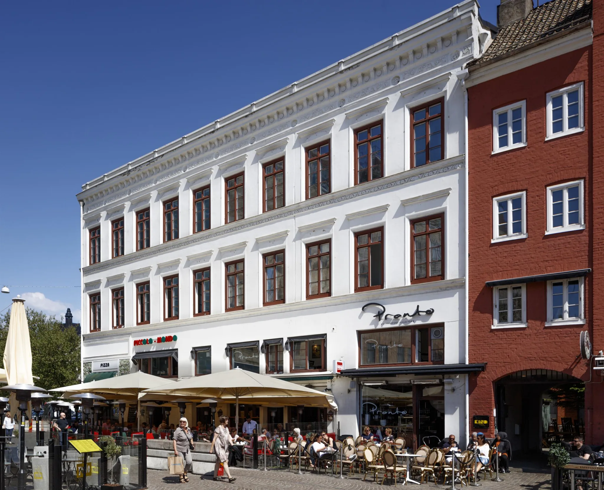 1. En vit byggnad med stora fönster och en uteservering med bord och stolar under parasoller på en solig dag.