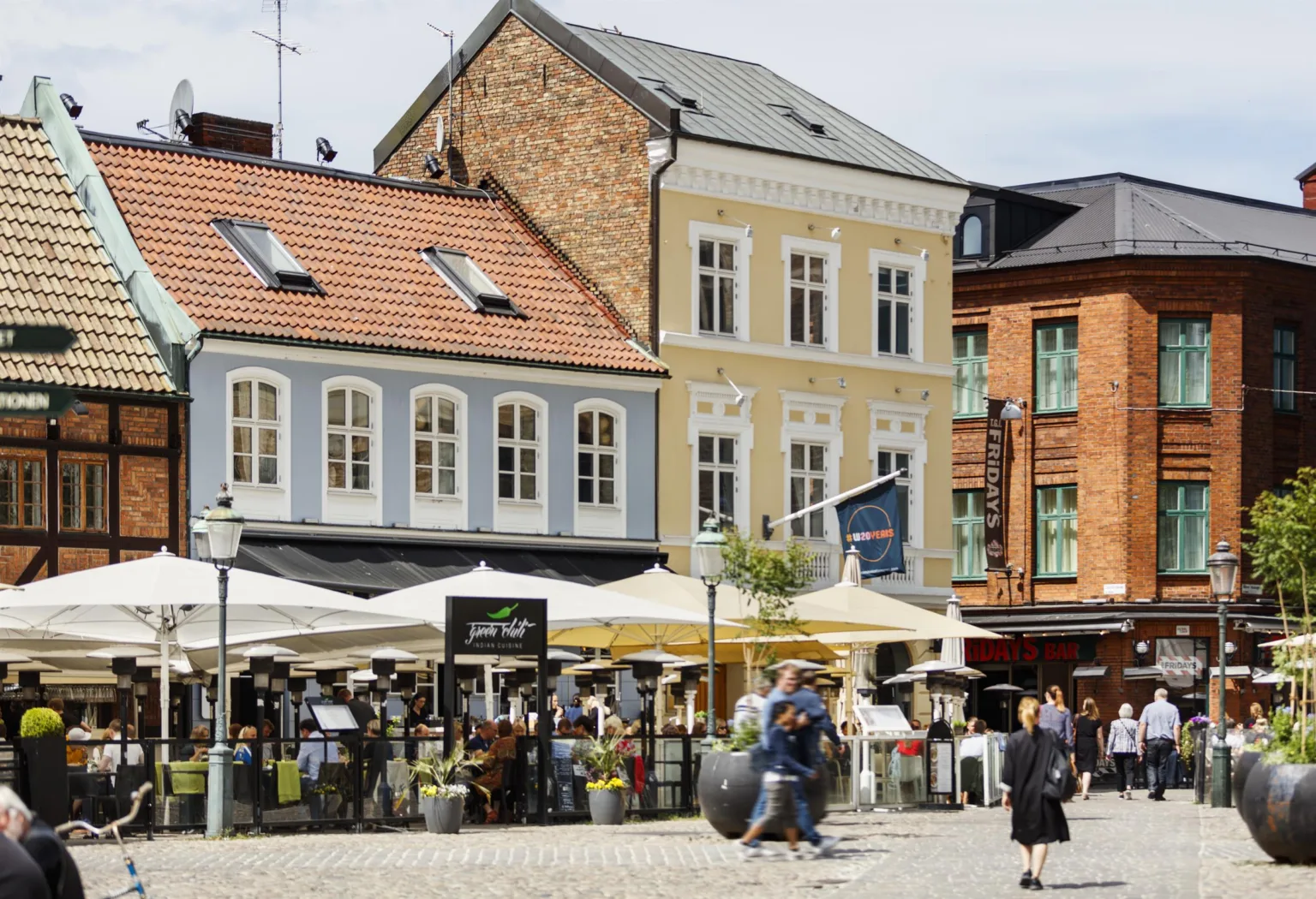 4. Ett torg med människor, uteserveringar under vita parasoller och färgglada byggnader i bakgrunden.