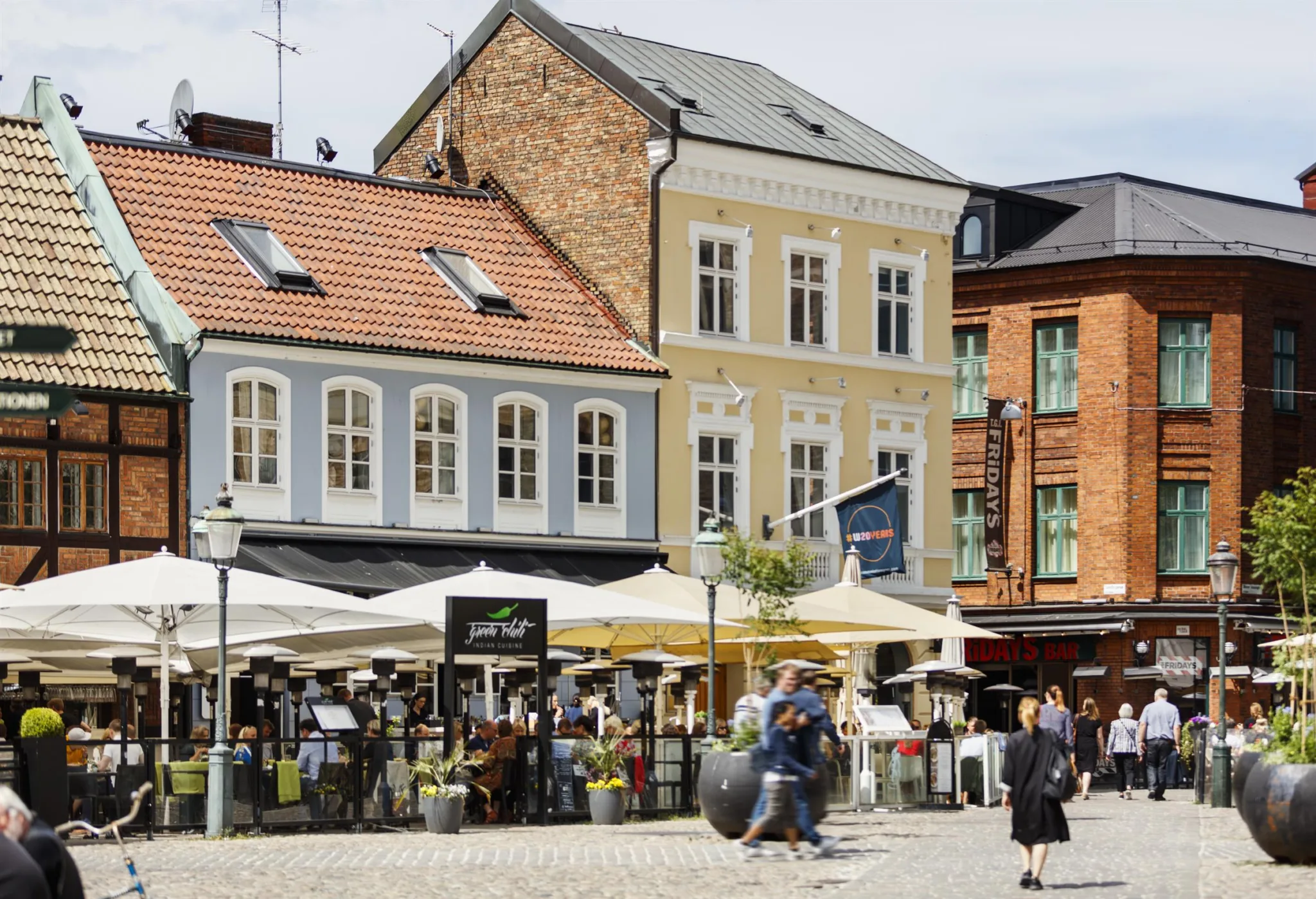 4. Ett torg med människor, uteserveringar under vita parasoller och färgglada byggnader i bakgrunden.