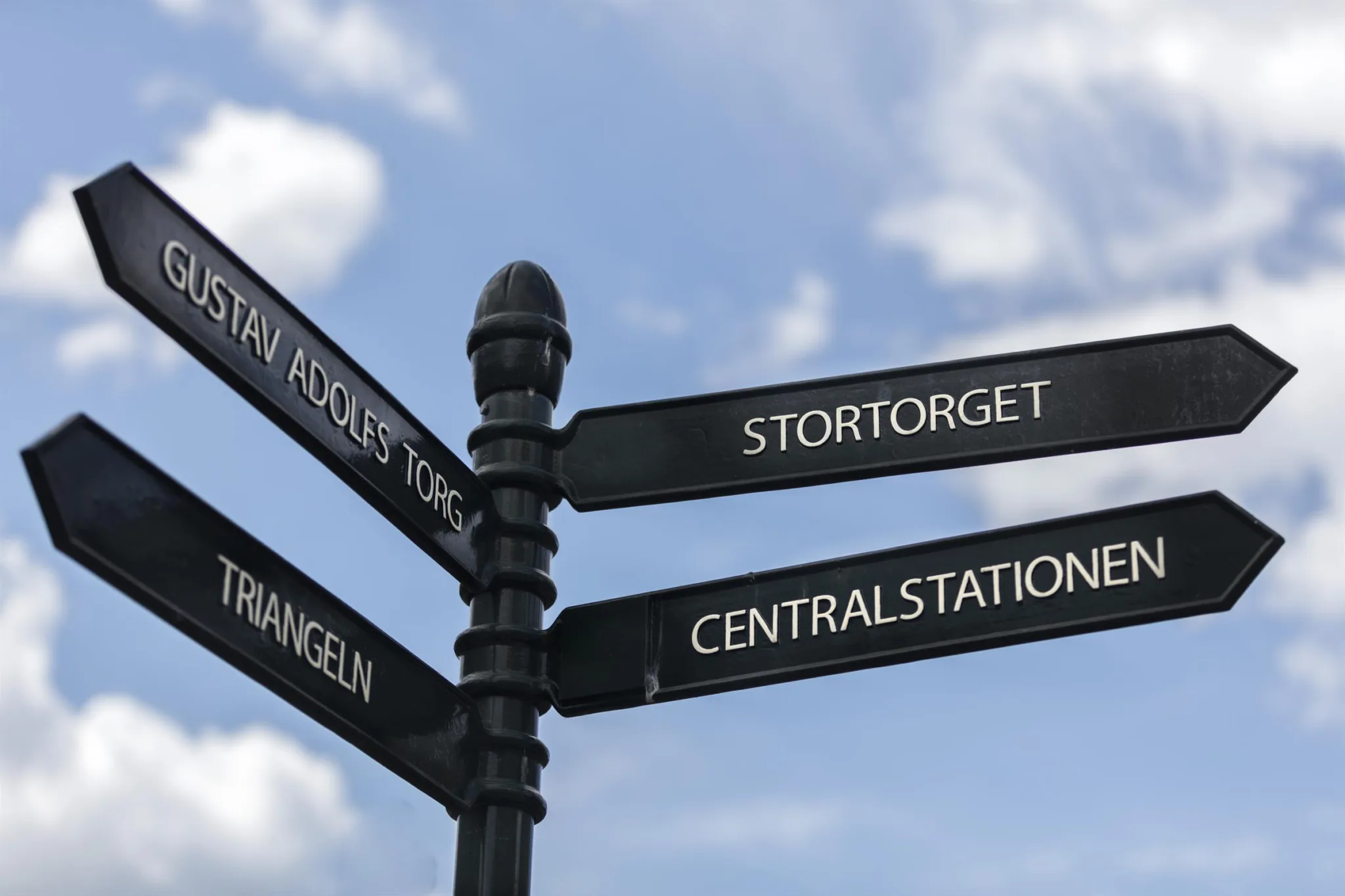 3. En svart vägskylt med fyra pilar som pekar mot "Stortorget", "Centralstationen", "Triangeln" och "Martin Andersens Torg", mot en bakgrund av blå himmel och vita moln.