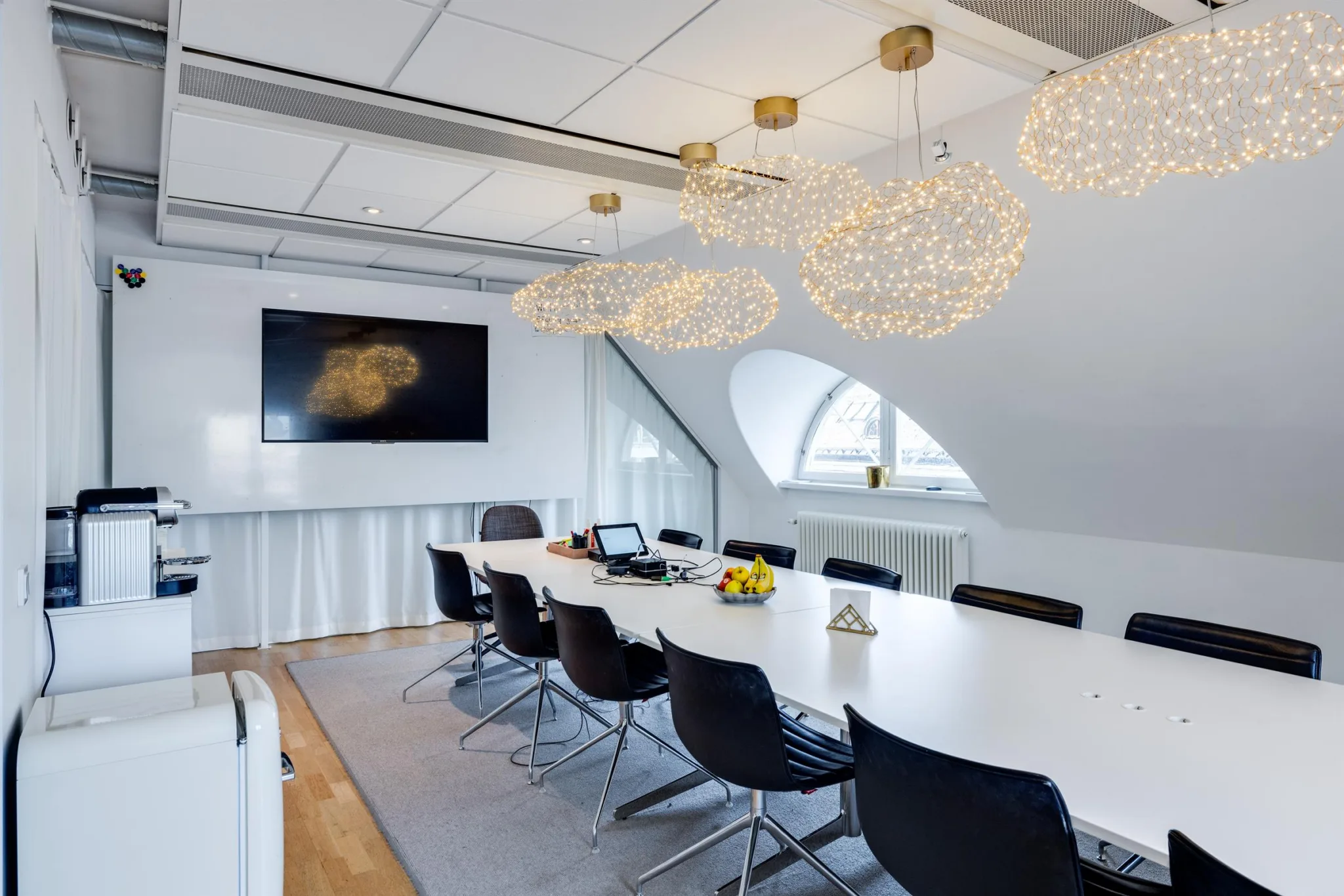 Ett mötesrum med lång konferensbord, svarta stolar, och dekorativa ljuskronor samt en väggmonterad TV.