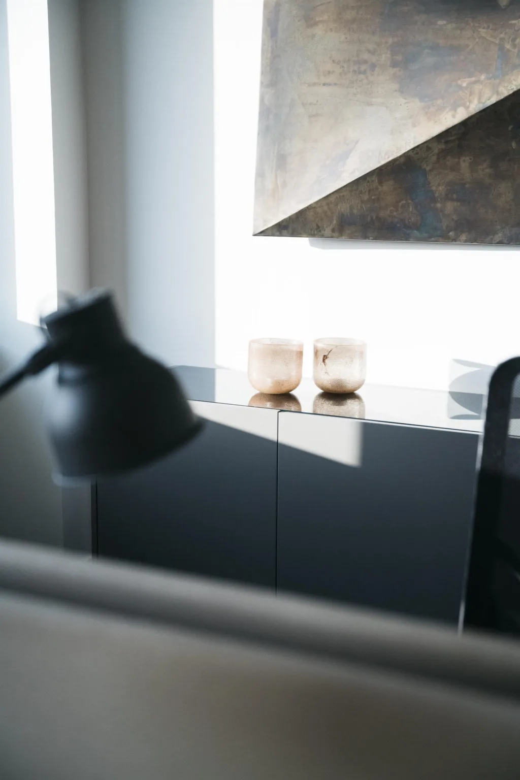 En bordslampa och två ljuslyktor på ett mörkt skåp, med geometrisk konst på väggen i bakgrunden.