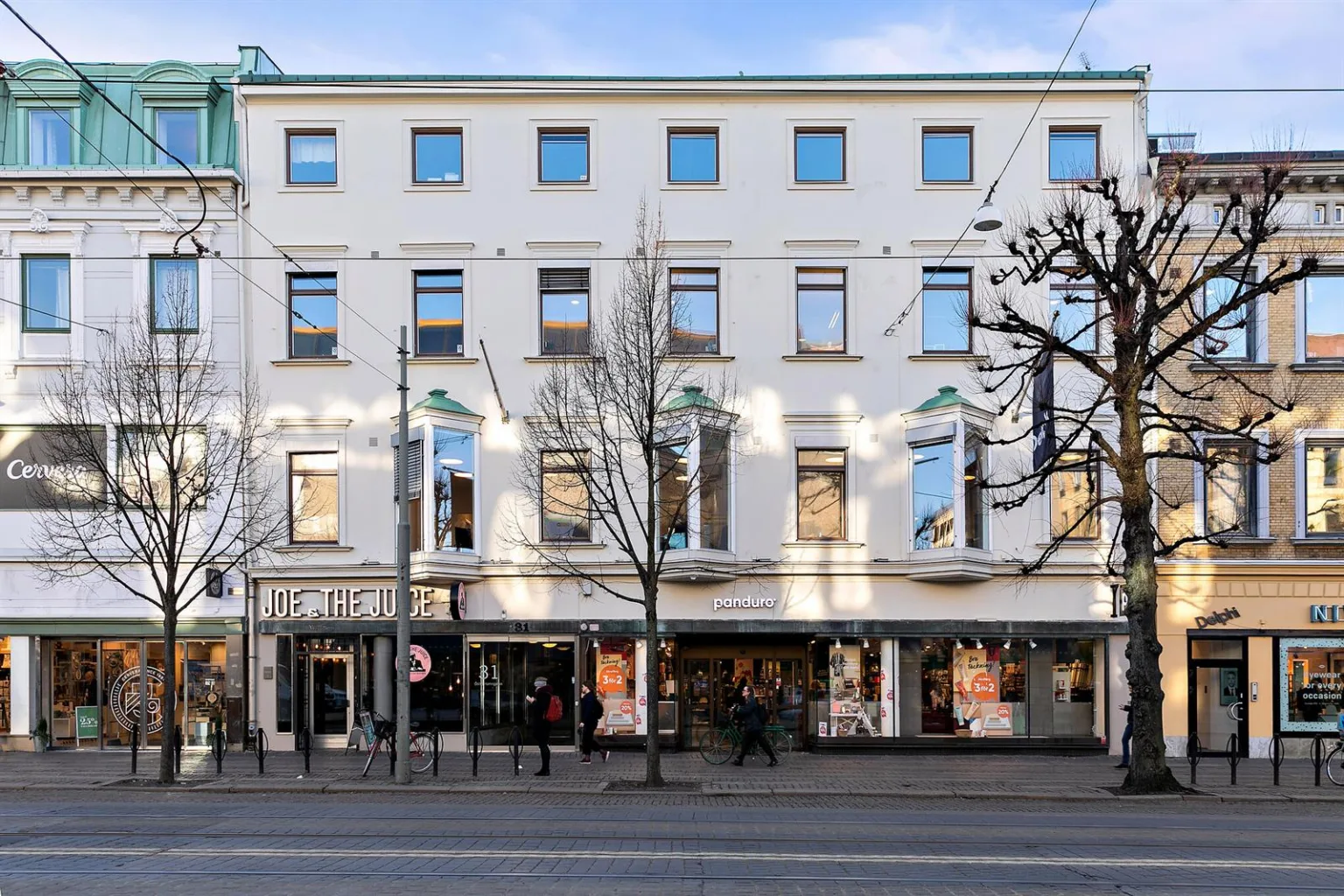 En vit byggnad med tre våningar och flera fönster; affärer och entréer finns på bottenvåningen, och träd står längs gatan framför.