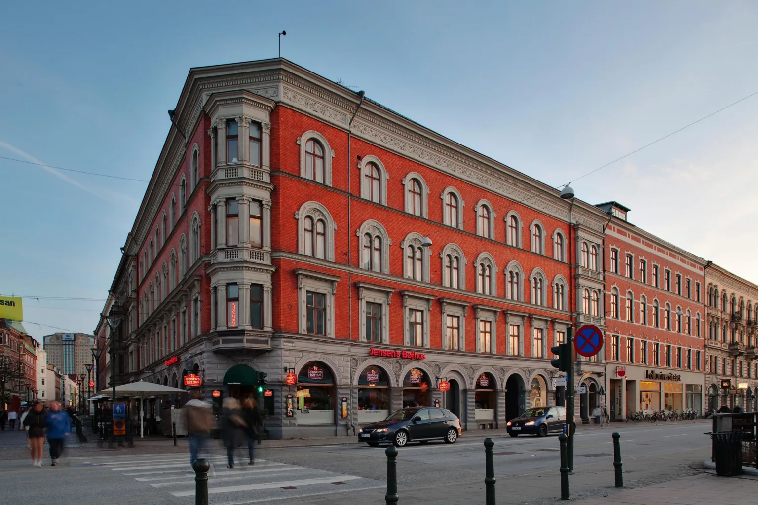 - En byggnad med röd fasad och stora fönster på en hörntomt, omgiven av trottoarer, lyktstolpar och parkerade bilar.