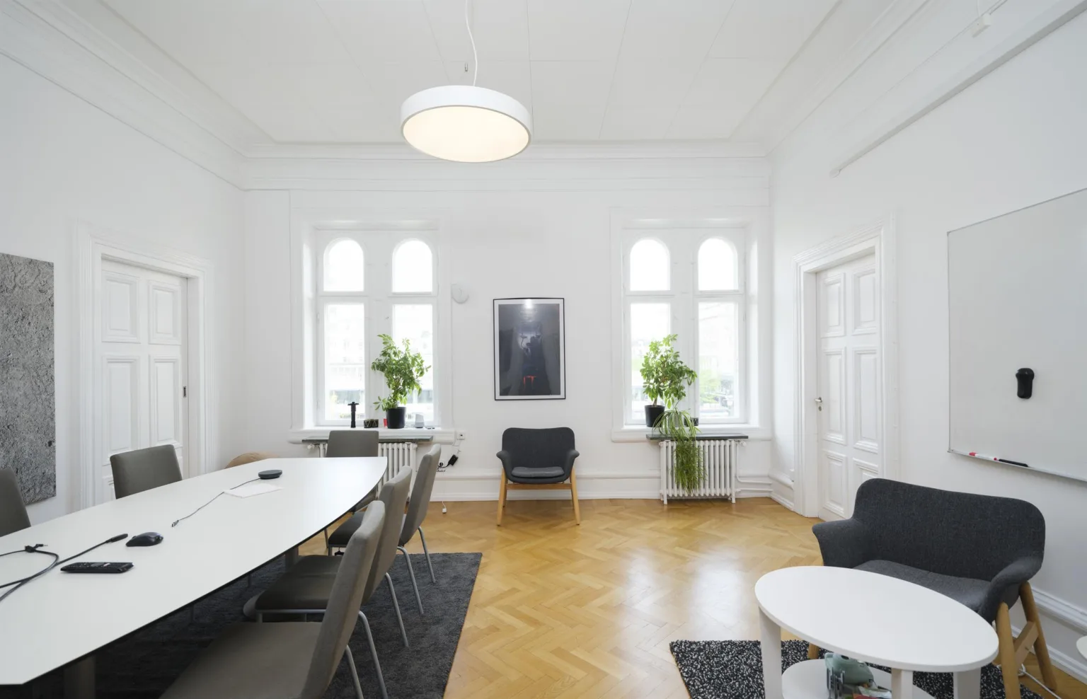 - Ett konferensrum med vita väggar, en lång bord med stolar, växter vid fönstren och en taklampa.