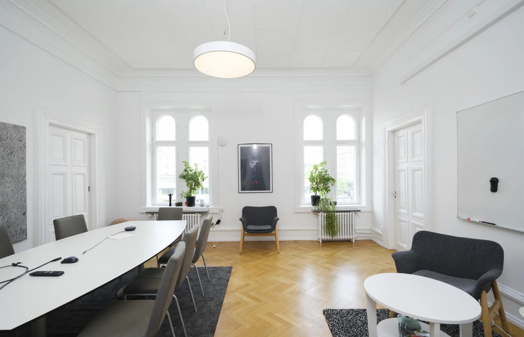 - Ett konferensrum med vita väggar, en lång bord med stolar, växter vid fönstren och en taklampa.