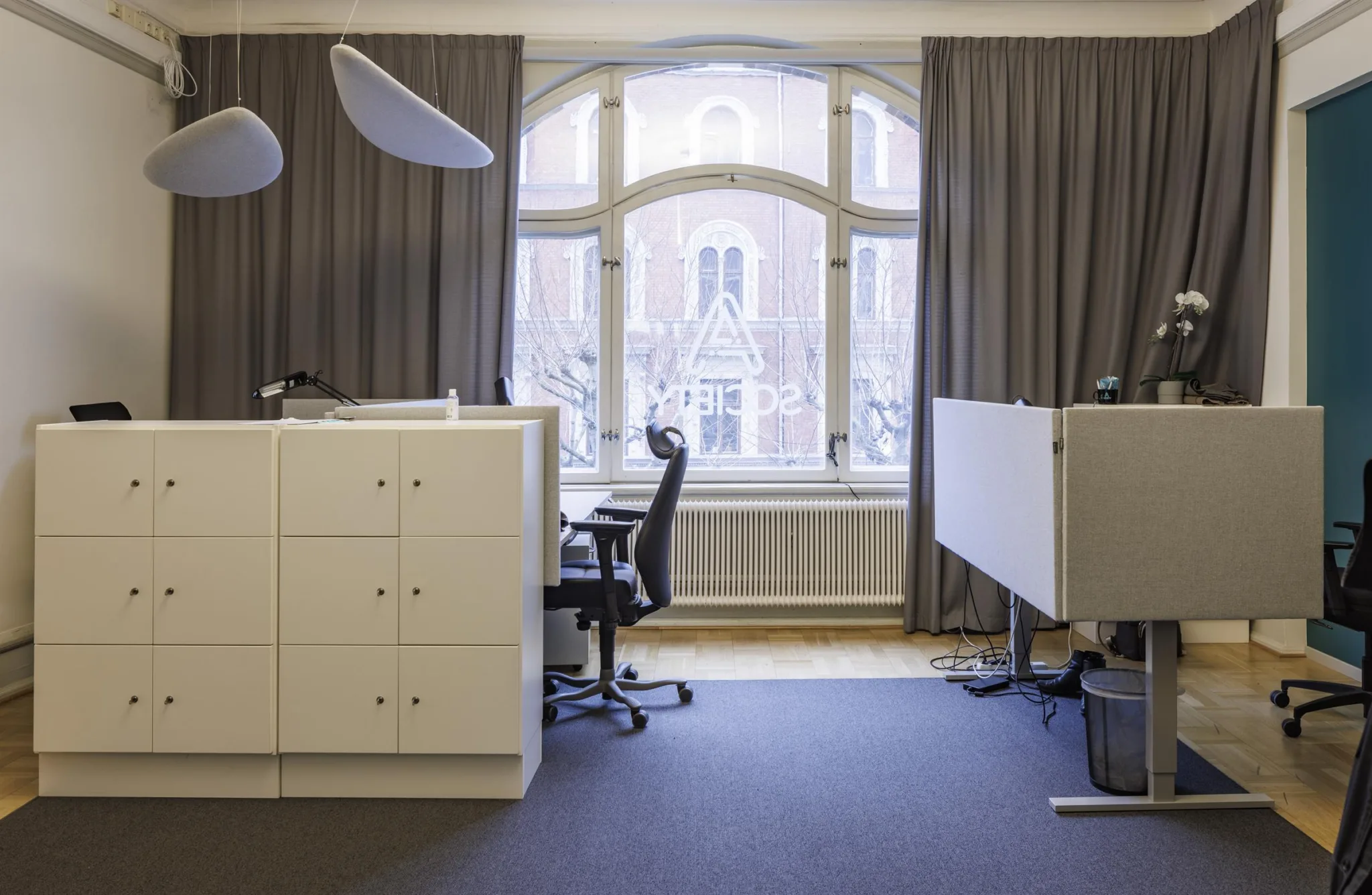 4. Ett kontor med två arbetsstationer, vita skrivbord, svarta stolar och stora fönster med gardiner som släpper in naturligt ljus.