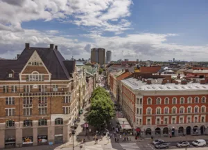 1. Vy över en stadsgata kantad av gamla tegelbyggnader med grönskande träd i mitten och höghus i bakgrunden under en delvis molnig himmel.