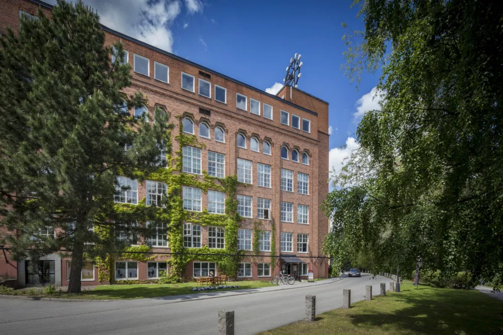 5. Tegelbyggnad med klätterväxter och stora fönster, omgiven av träd och gräsmatta.