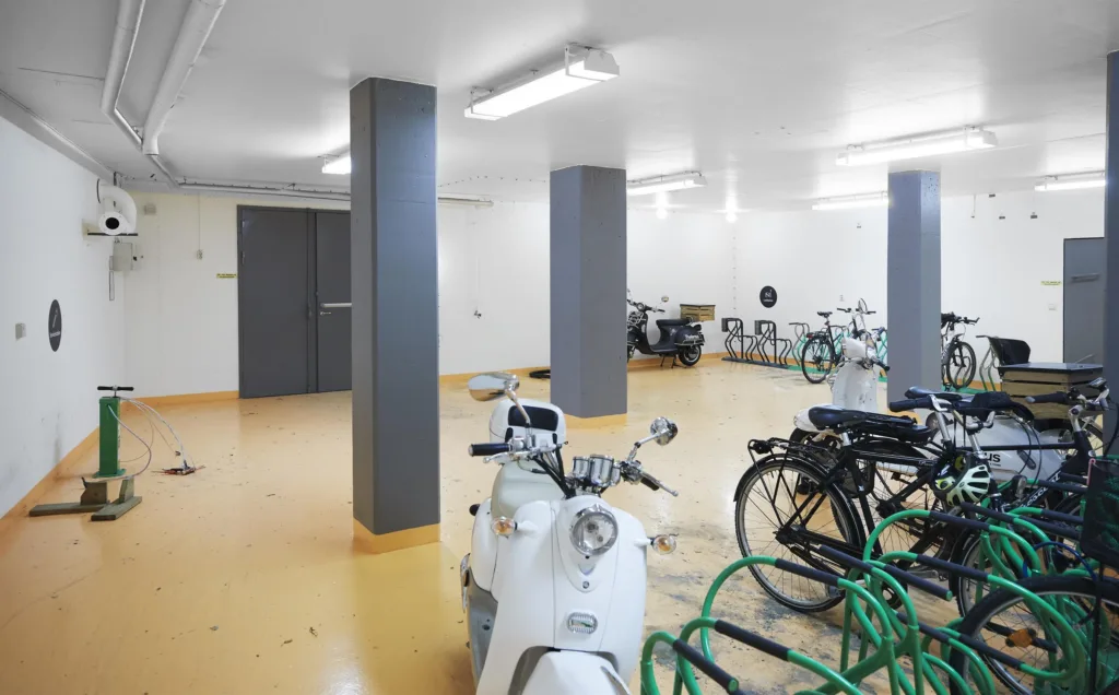 En ljus cykel- och mopedparkering med vita väggar och pelare, flera cyklar och mopeder står parkerade.