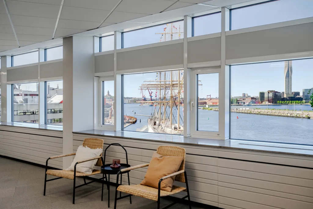 4. Två fåtöljer och ett litet bord placerade framför stora fönster med utsikt över en hamn och segelbåtar.