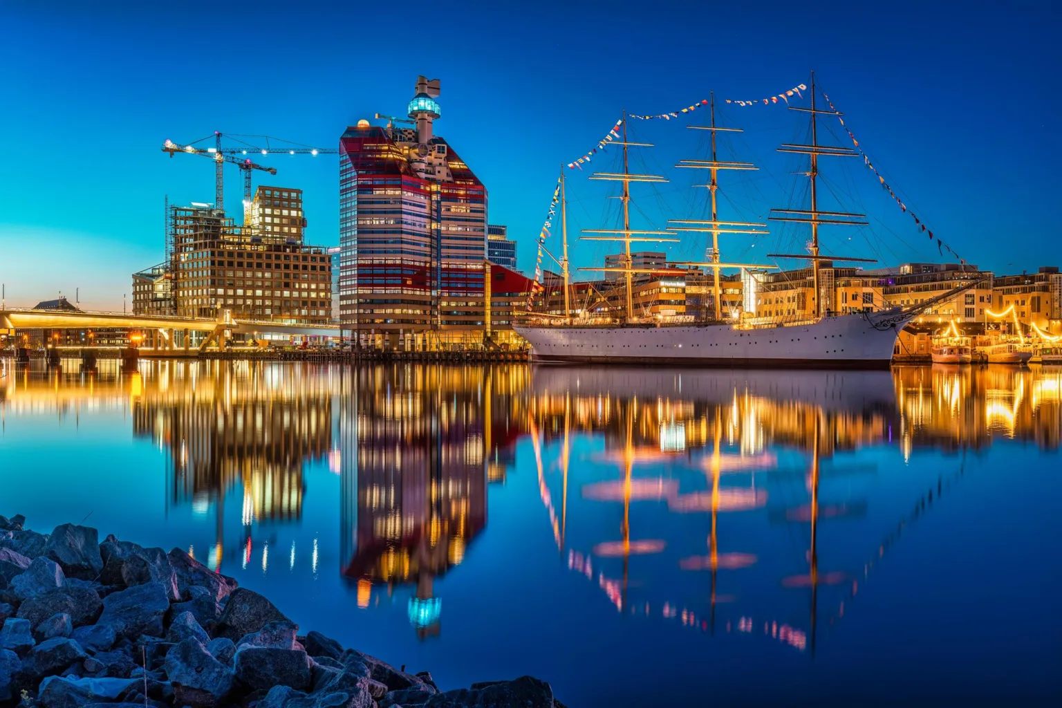 En nattvy av Göteborg med den upplysta byggnaden "Läppstiftet" och ett segelfartyg som reflekteras i vattnet.