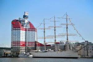 Ett stort segelfartyg med flera master nära en rödvit byggnad med glasfasad.