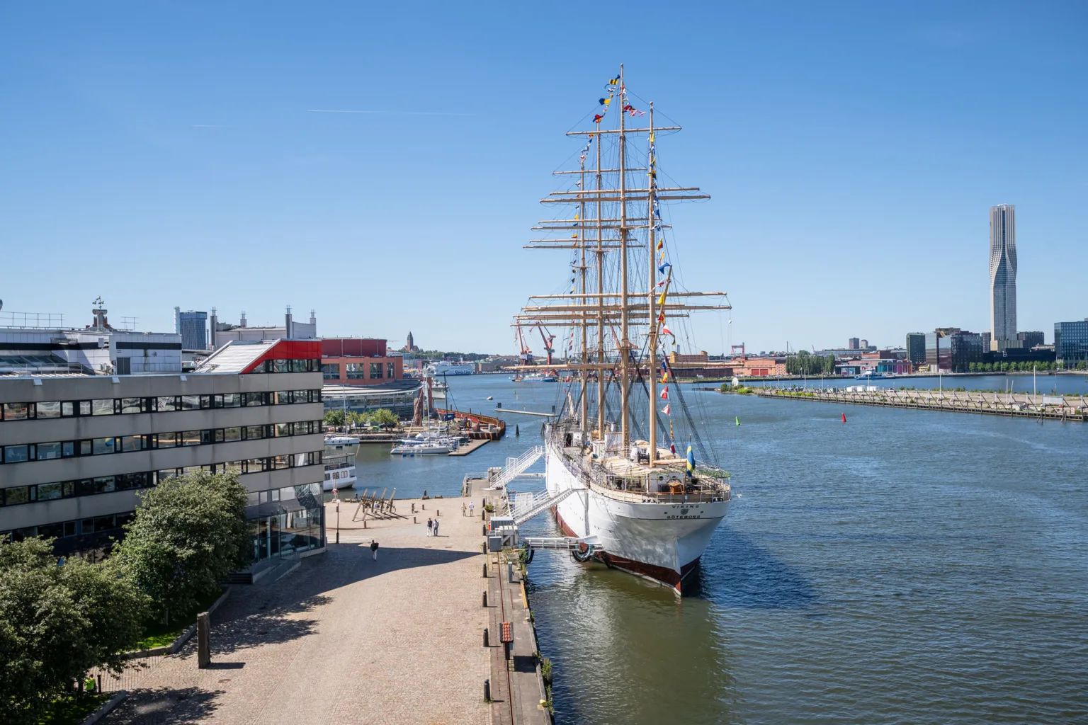 En vit segelbåt förtöjd vid en kaj i en stadsmiljö, med byggnader och blå himmel i bakgrunden.