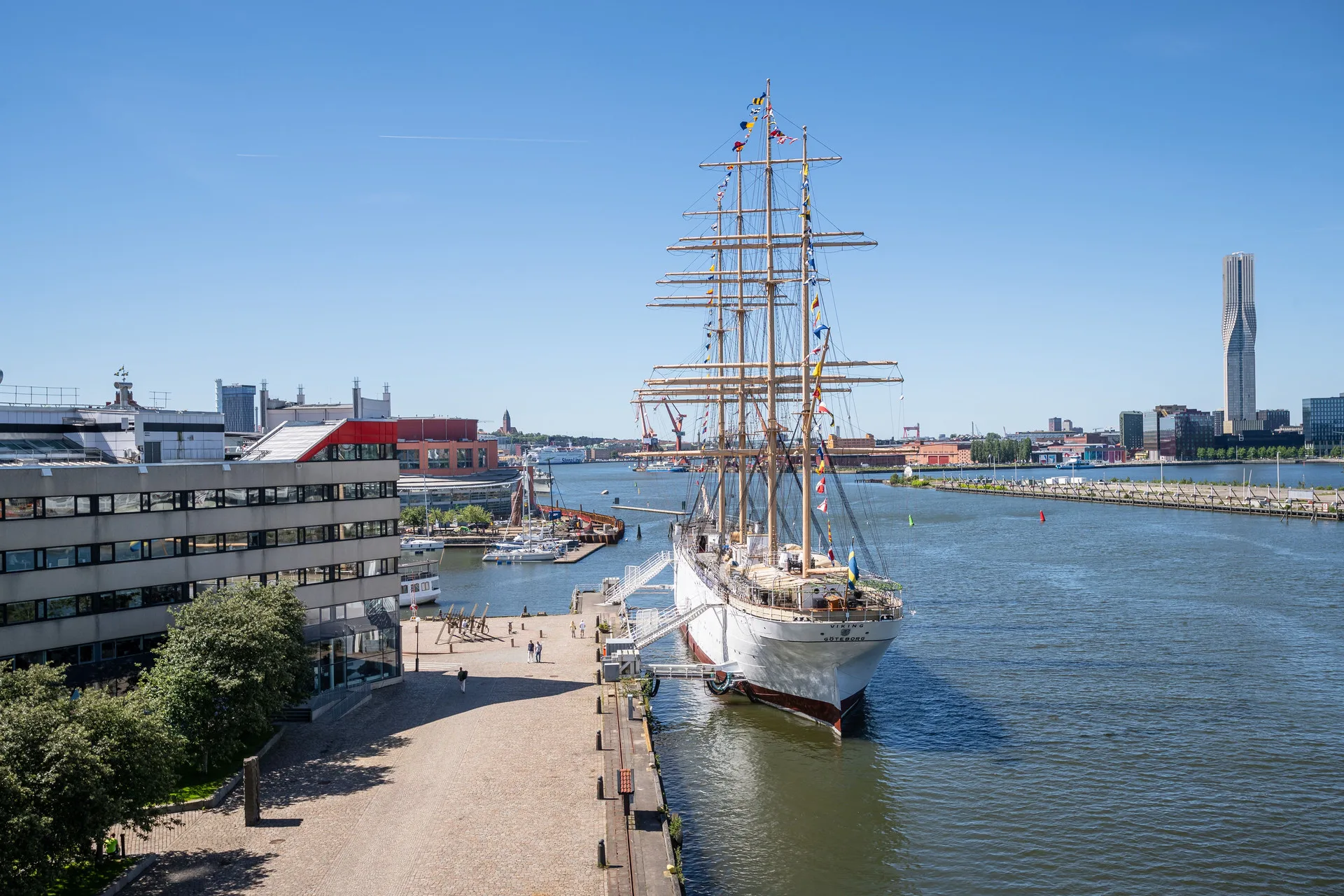 En vit segelbåt förtöjd vid en kaj i en stadsmiljö, med byggnader och blå himmel i bakgrunden.