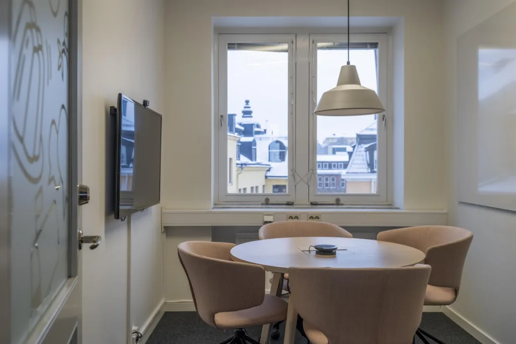 5. Ett mötesrum med ett runt bord, stolar, en väggmonterad skärm och fönster med utsikt över stadens tak.