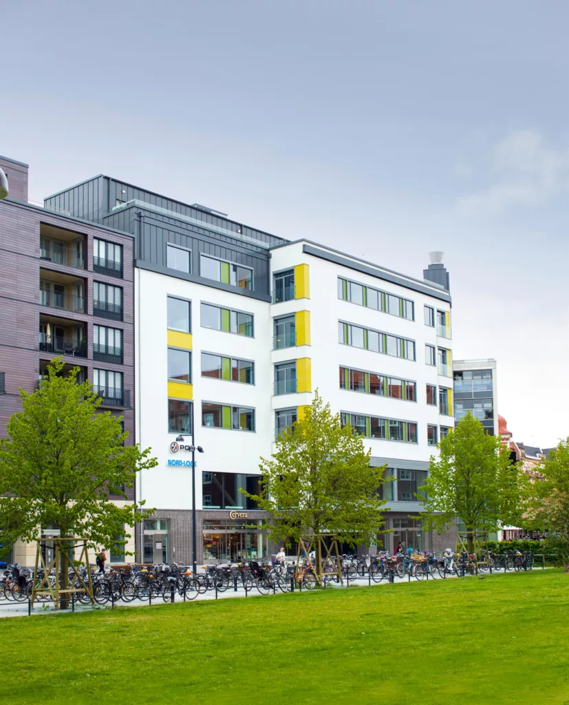 En modern byggnad med vita och färgade fasader, omgiven av gröna träd och en gräsmatta.