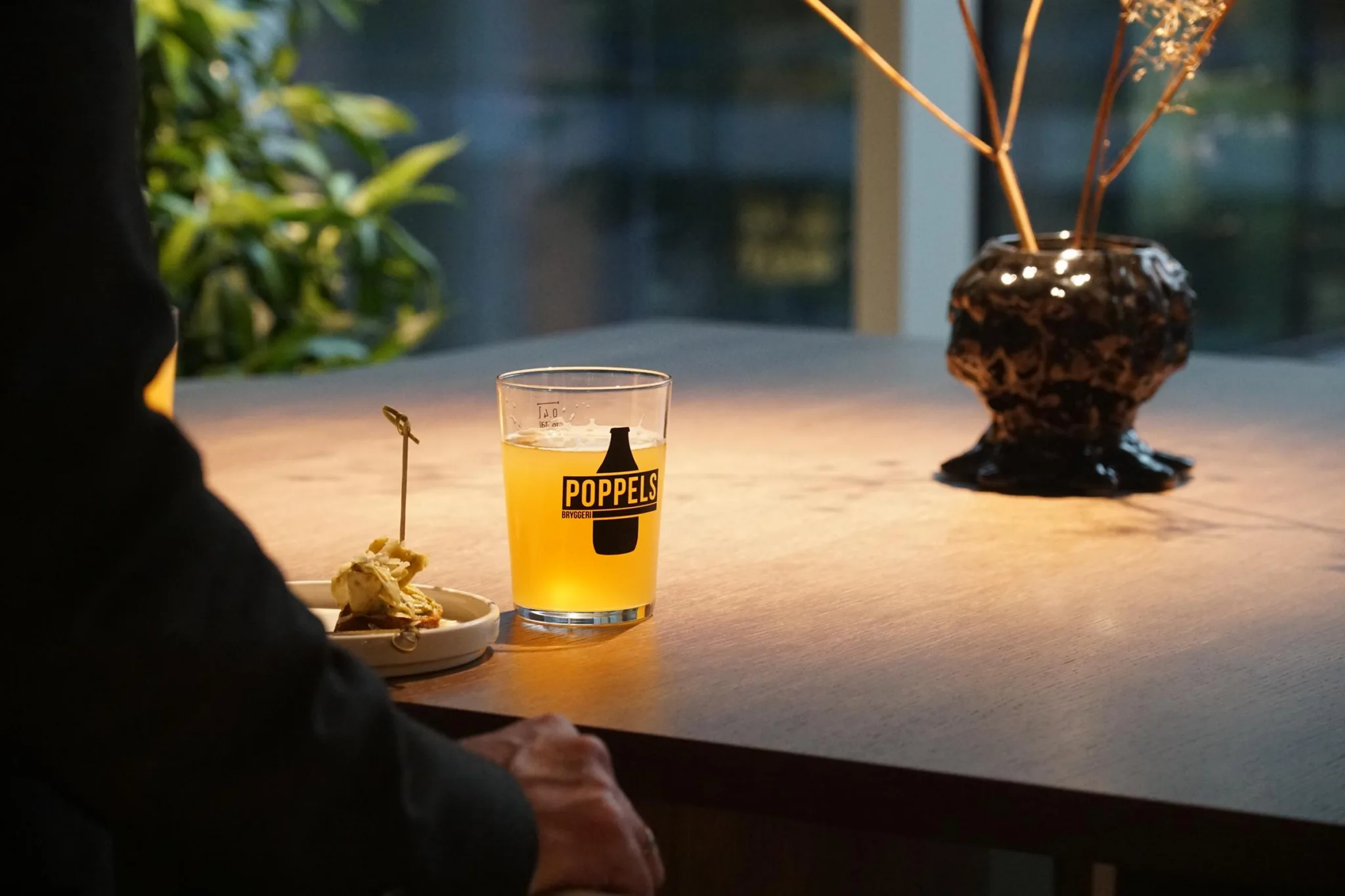 2. Ett glas med gul dryck på ett bord, bredvid en tallrik med en bit mat, i en miljö med växter och mjuk belysning.