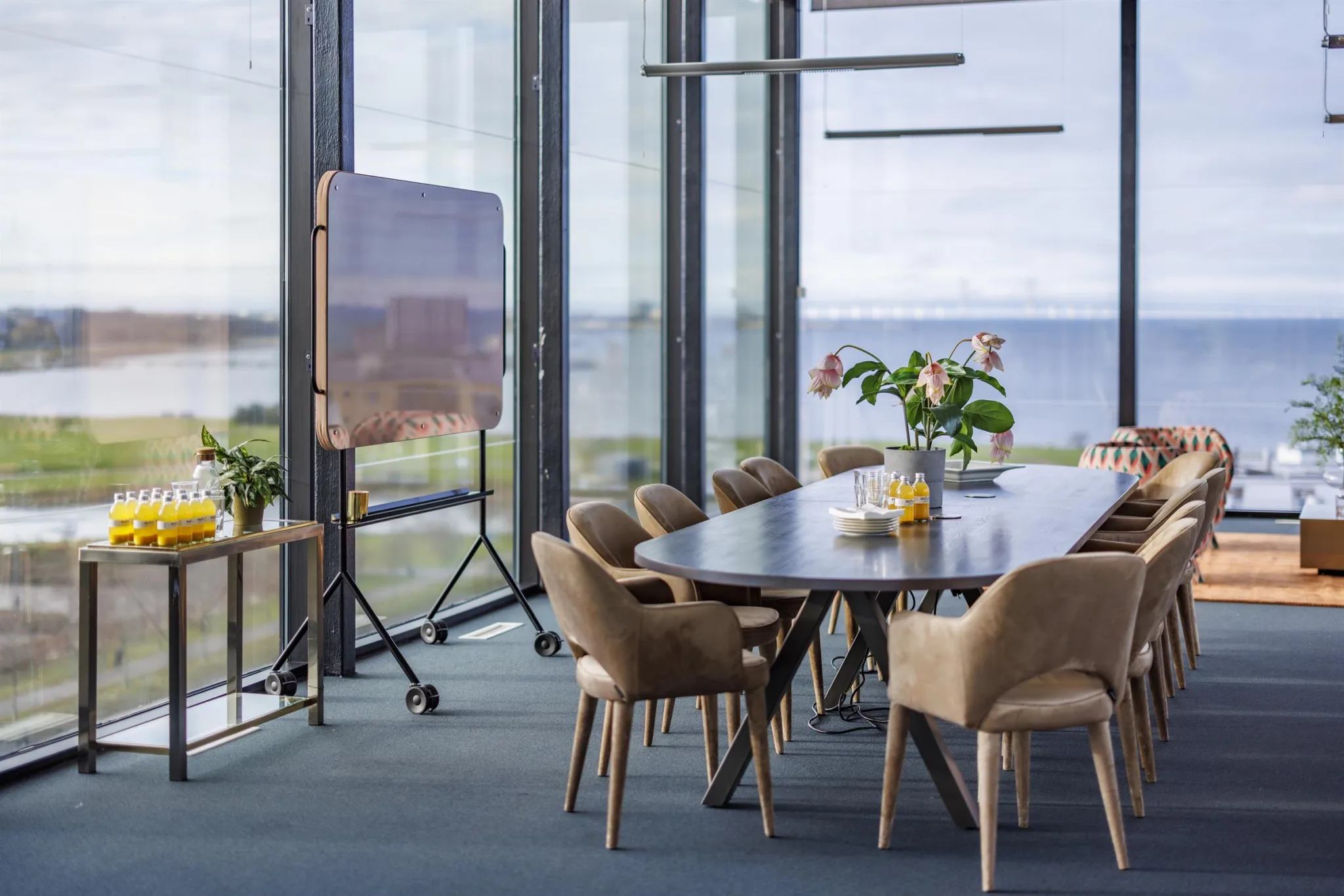 2. Konferensrum med stort bord, beige stolar, blommor och utsikt över vatten genom stora fönster.