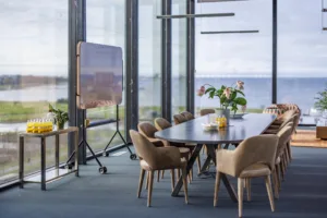 2. Konferensrum med stort bord, beige stolar, blommor och utsikt över vatten genom stora fönster.