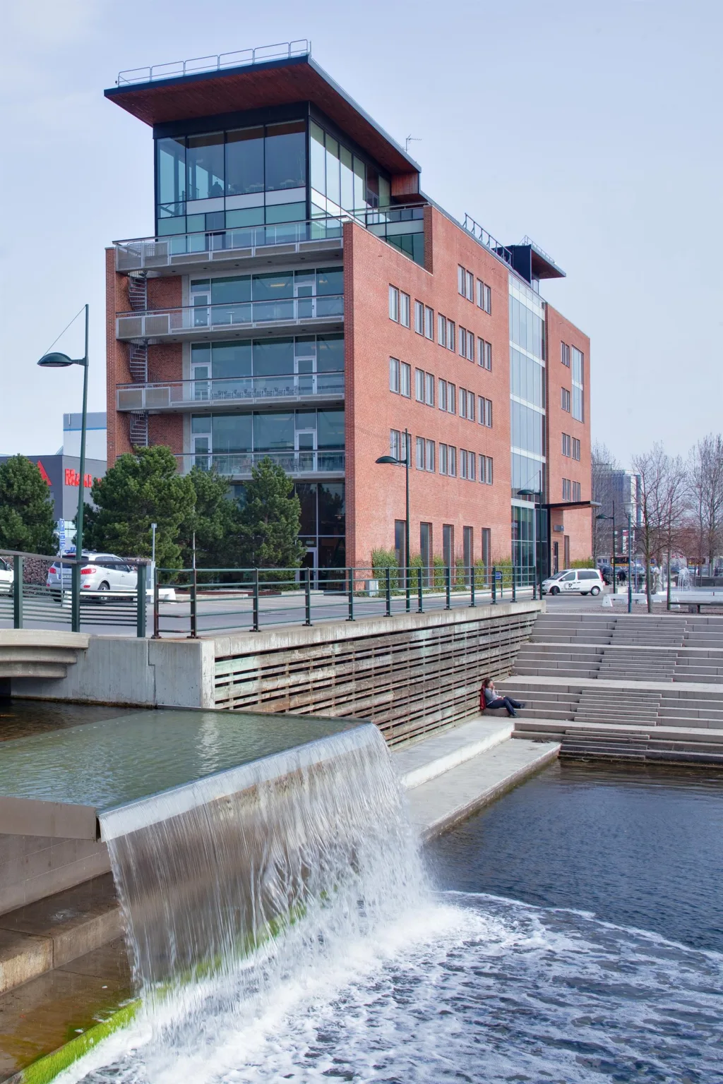 Ett modernt tegelhus med flera våningar, stora glasfönster och en liten forsande vattenkanal framför byggnaden.