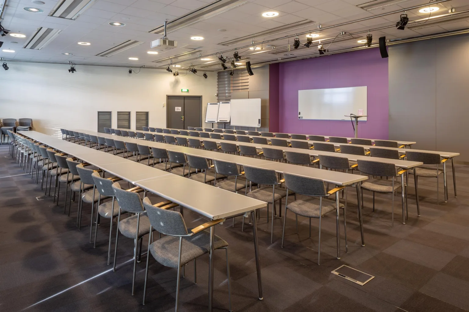 Ett konferensrum med långbord och stolar, en lila vägg med två whiteboards och en projektorskärm.