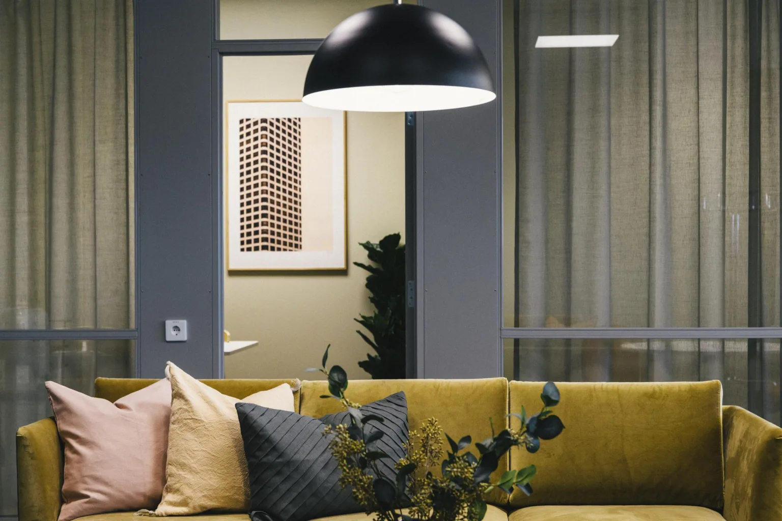 En gul soffa med färgglada kuddar, en svart taklampa och växtdekorationer i en modern miljö.