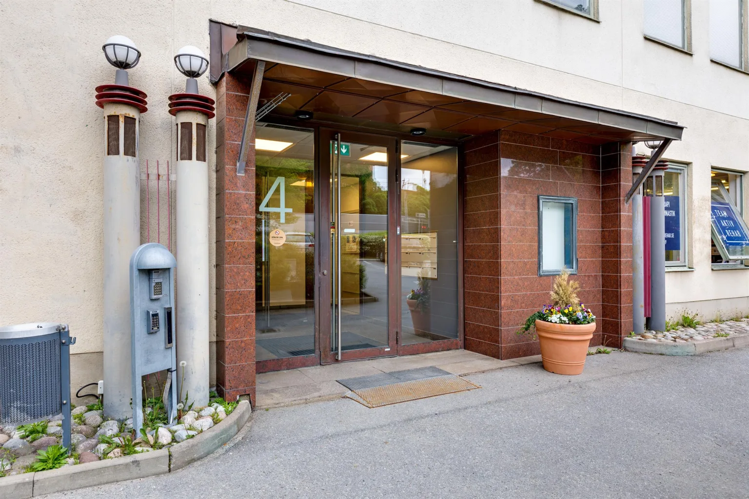 2. Ingång till en byggnad med glasdörrar, röd tegelvägg och en krukväxt bredvid.